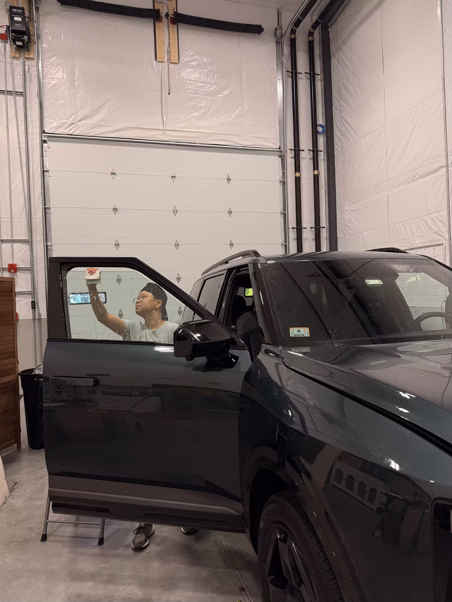 Technician applying tint film to the open front driver door window of a dark SUV in a garage bay.
