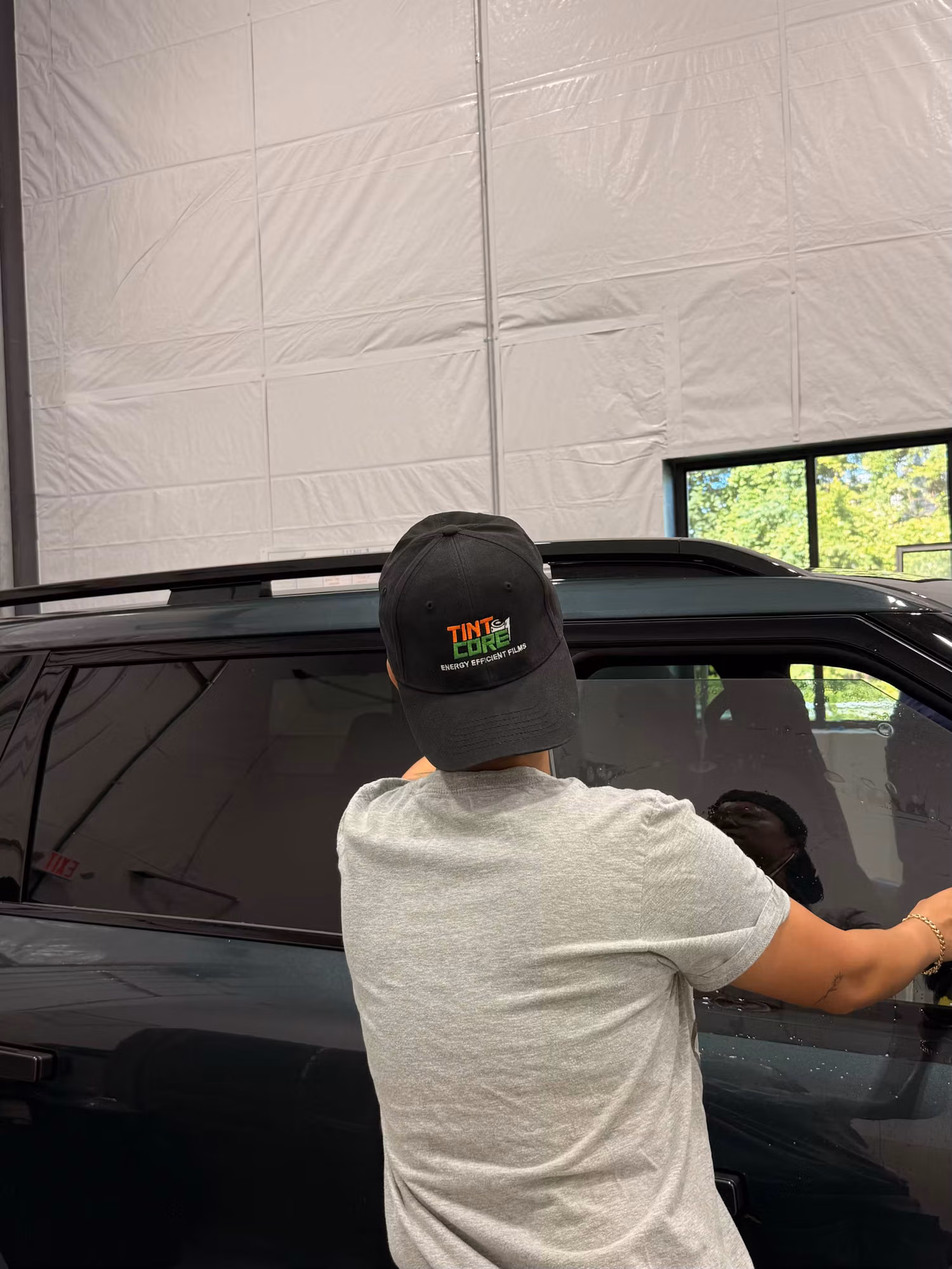 Installer in a branded cap smoothing tint film onto the rear side window of a dark SUV.