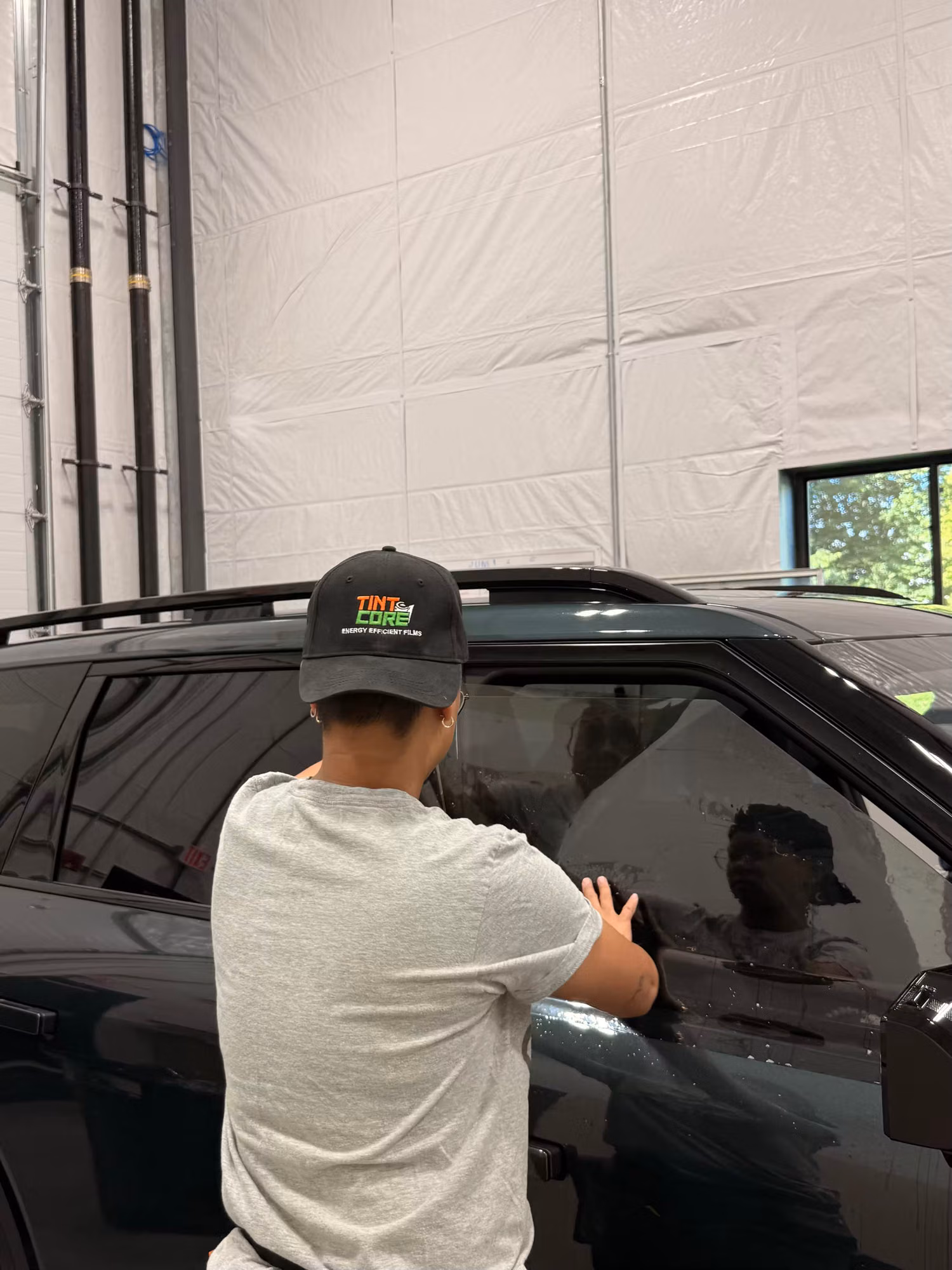 Technician squeegeeing tint film onto a darkened rear door window of an SUV inside a shop bay.