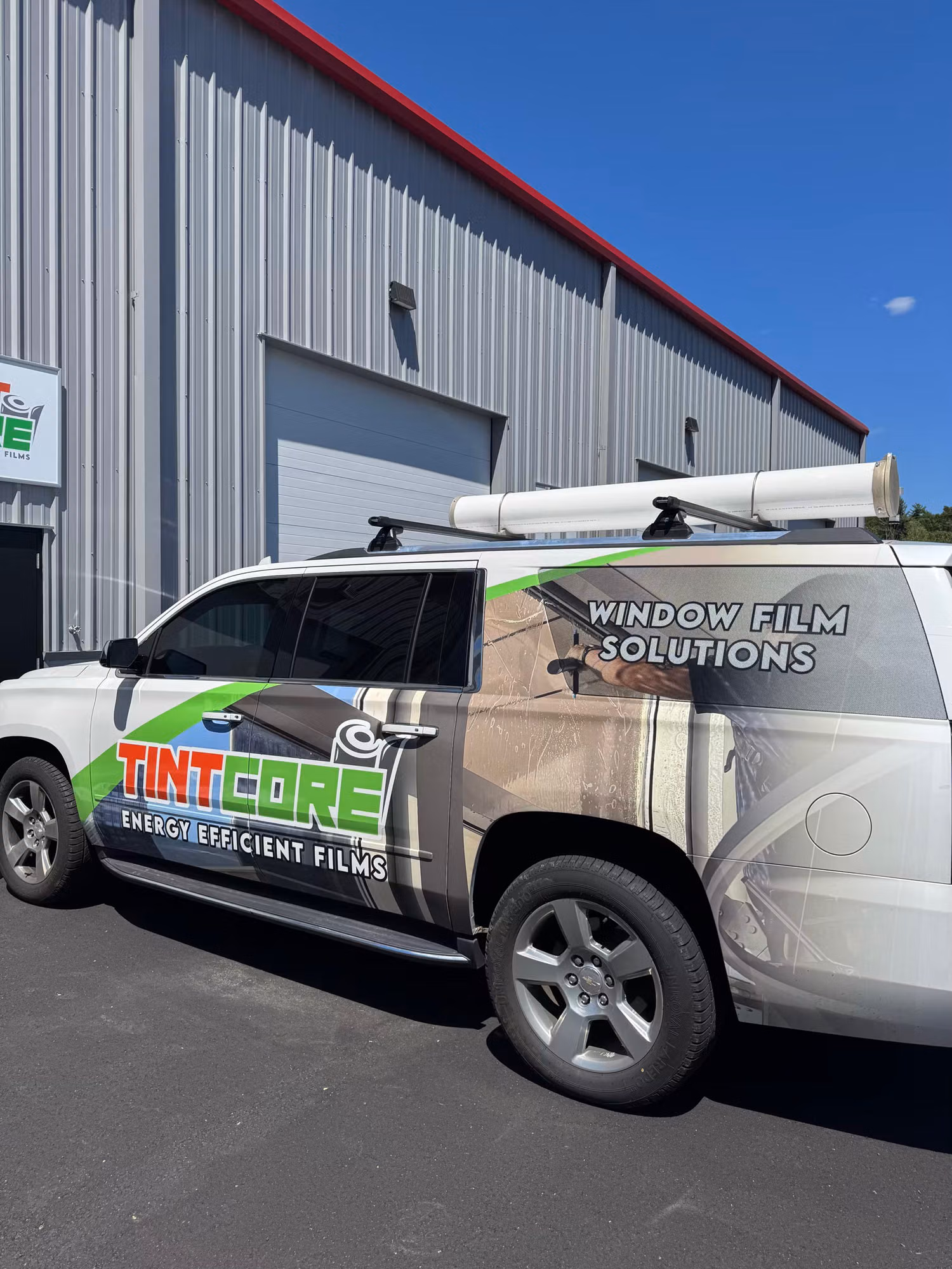 Wrapped service SUV with a roof-mounted film tube parked beside a metal warehouse under a clear sky.