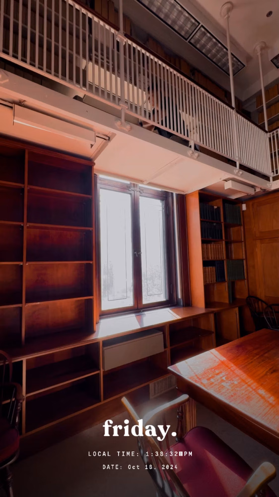 Tall window in a two-story library reading room with empty wooden bookshelves and a mezzanine balcony.
