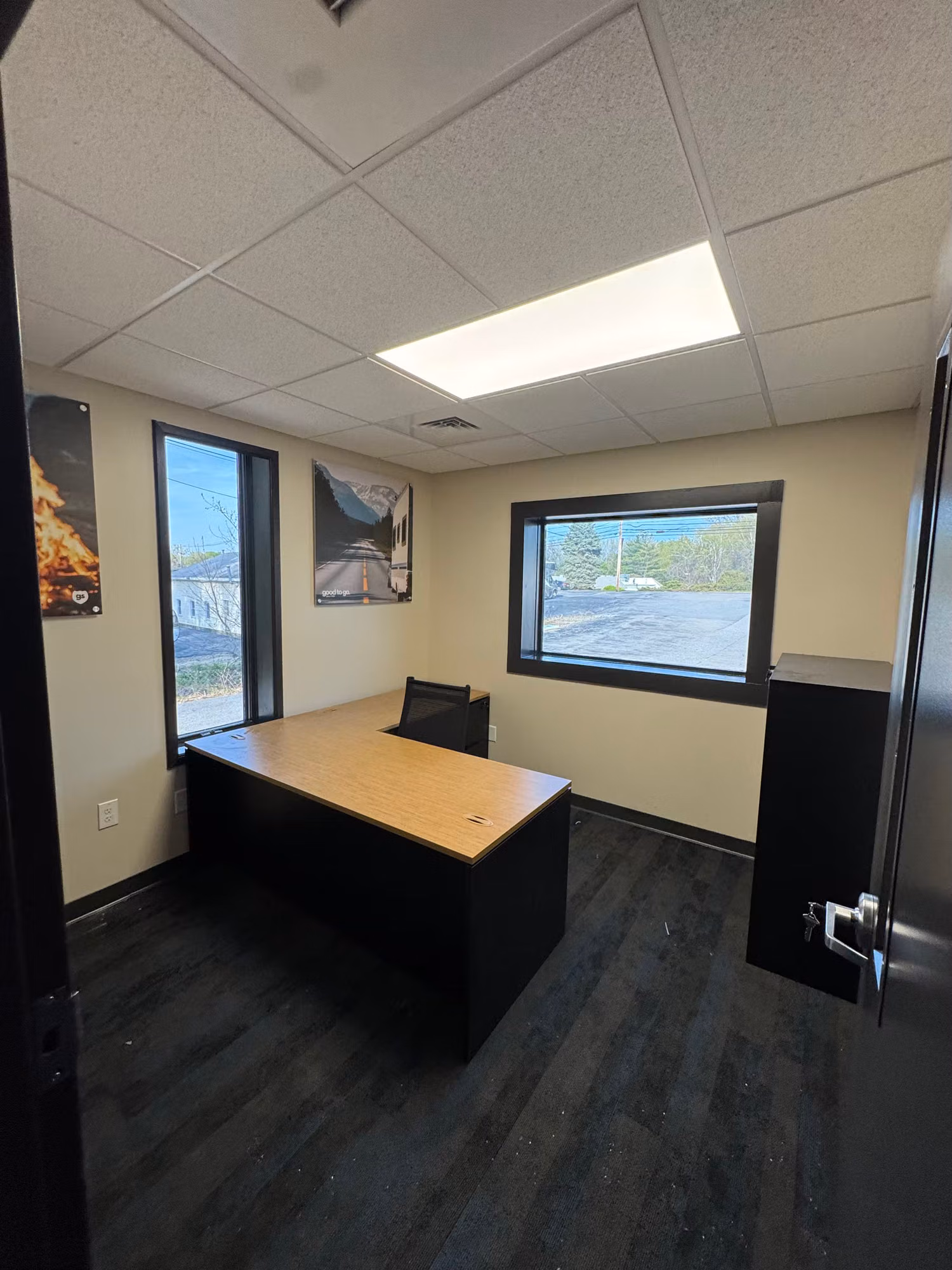 Small commercial office with perimeter windows around an L-shaped desk and dark flooring.