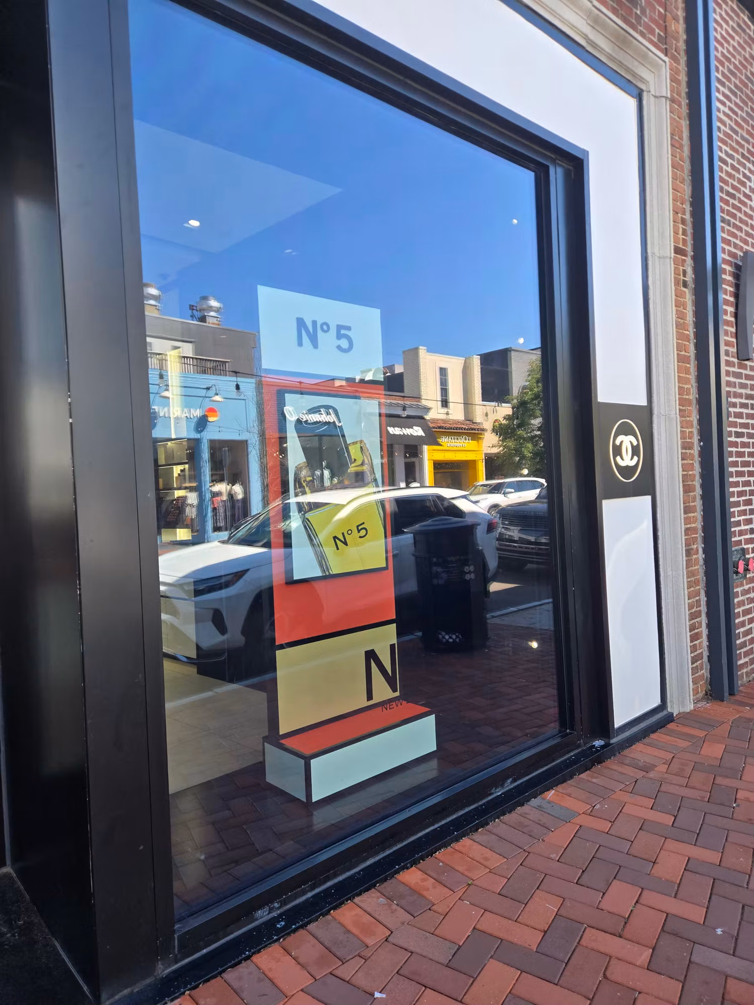 Large black-framed retail storefront window with clear protective film and street reflections along a brick sidewalk.