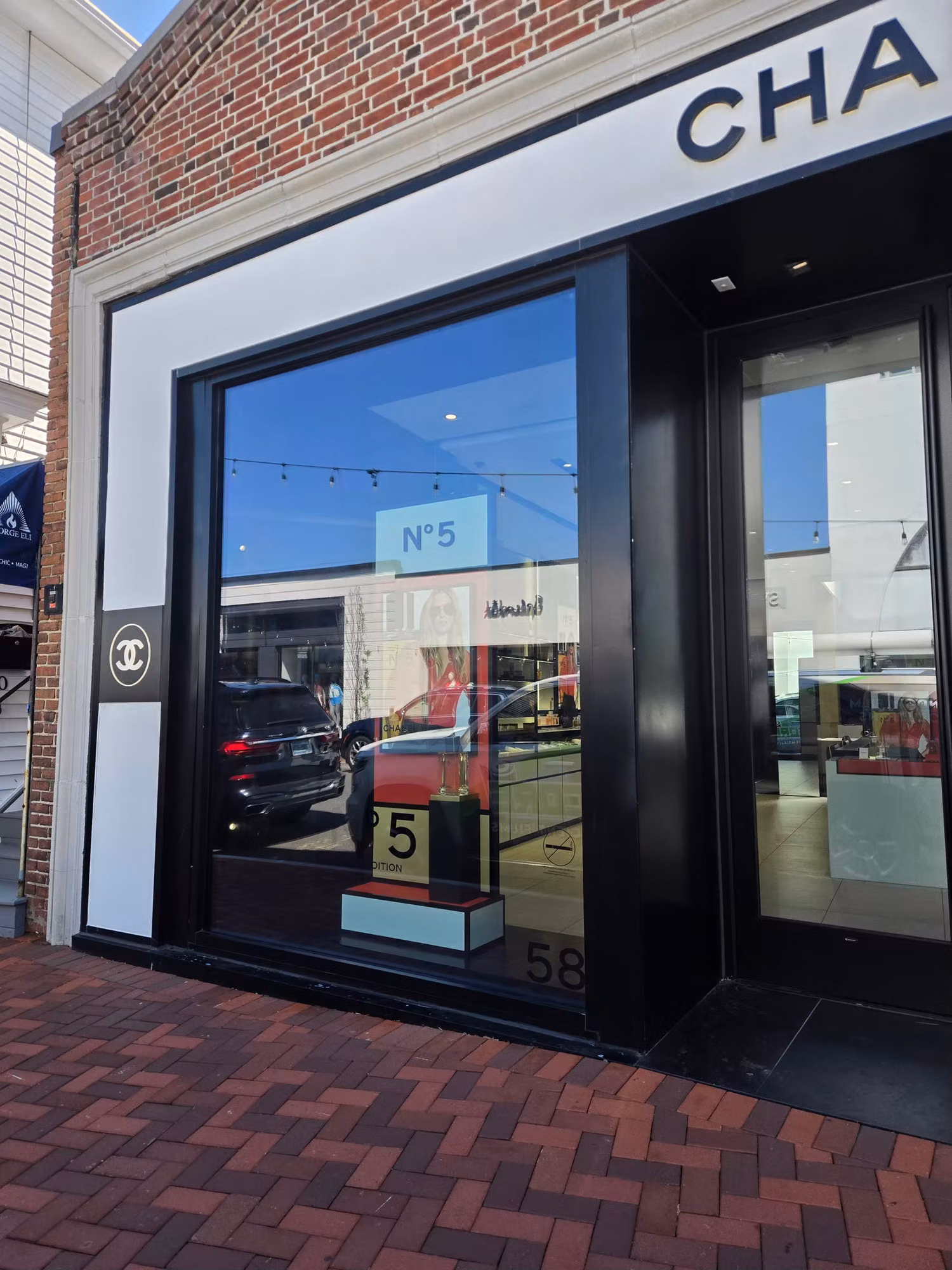 Wide retail storefront display window in a black frame with clear protective film reflecting the street and brick walkway.