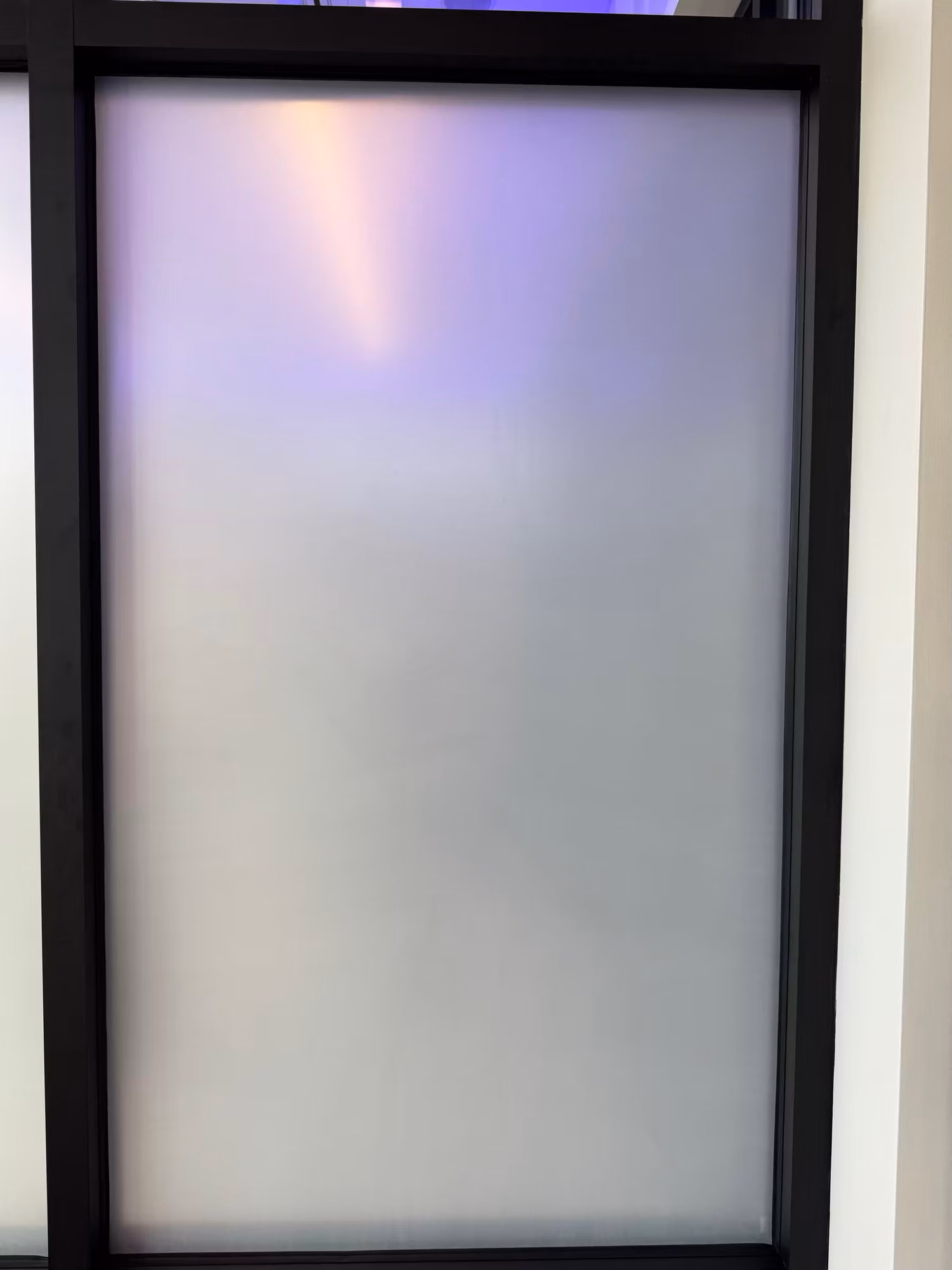 Close-up of a black-framed glass partition panel covered with frosted privacy film for an opaque, light-diffusing finish.