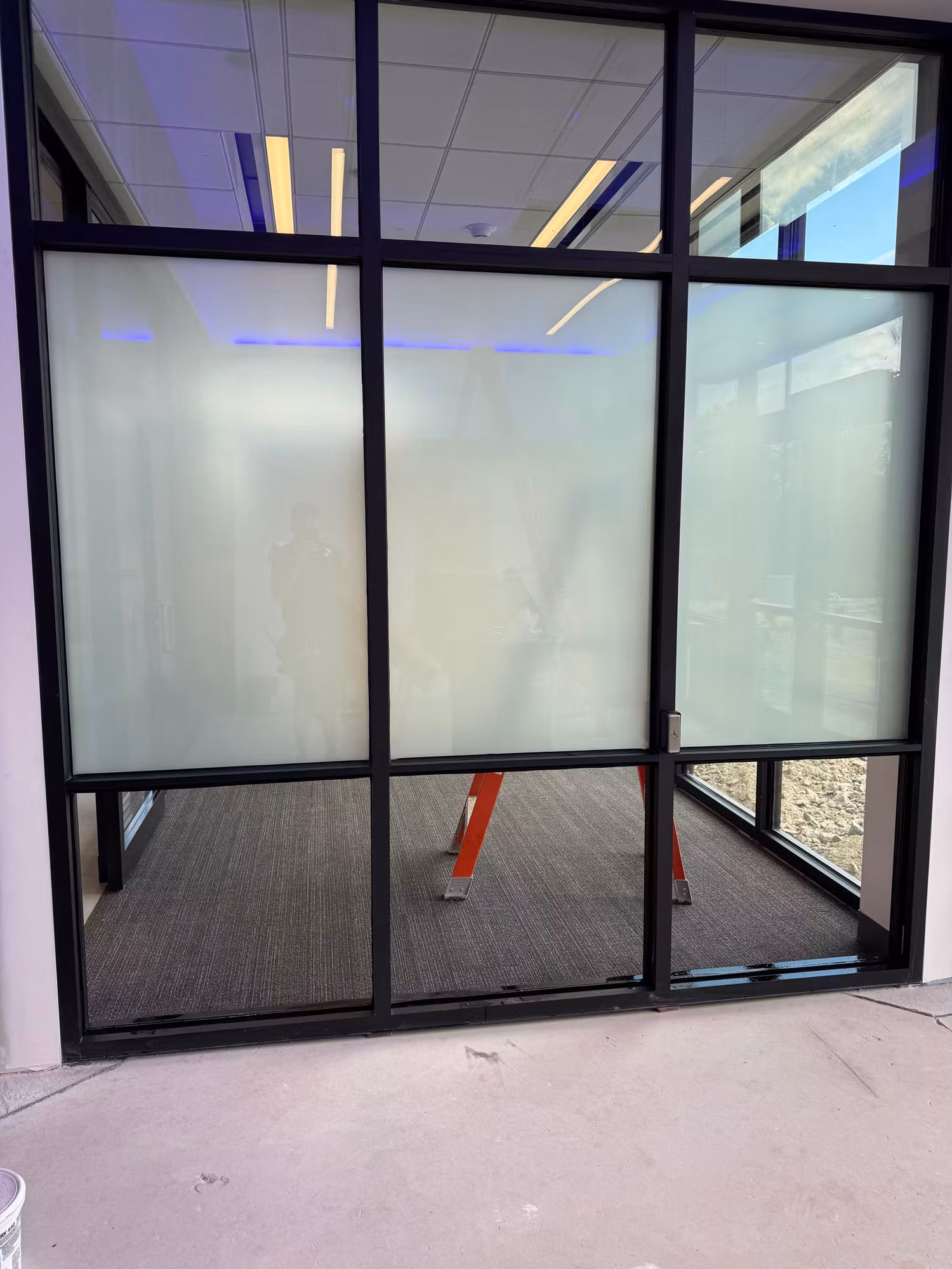 Interior glass partition with framed doors showing frosted privacy film banding across the panels during installation.