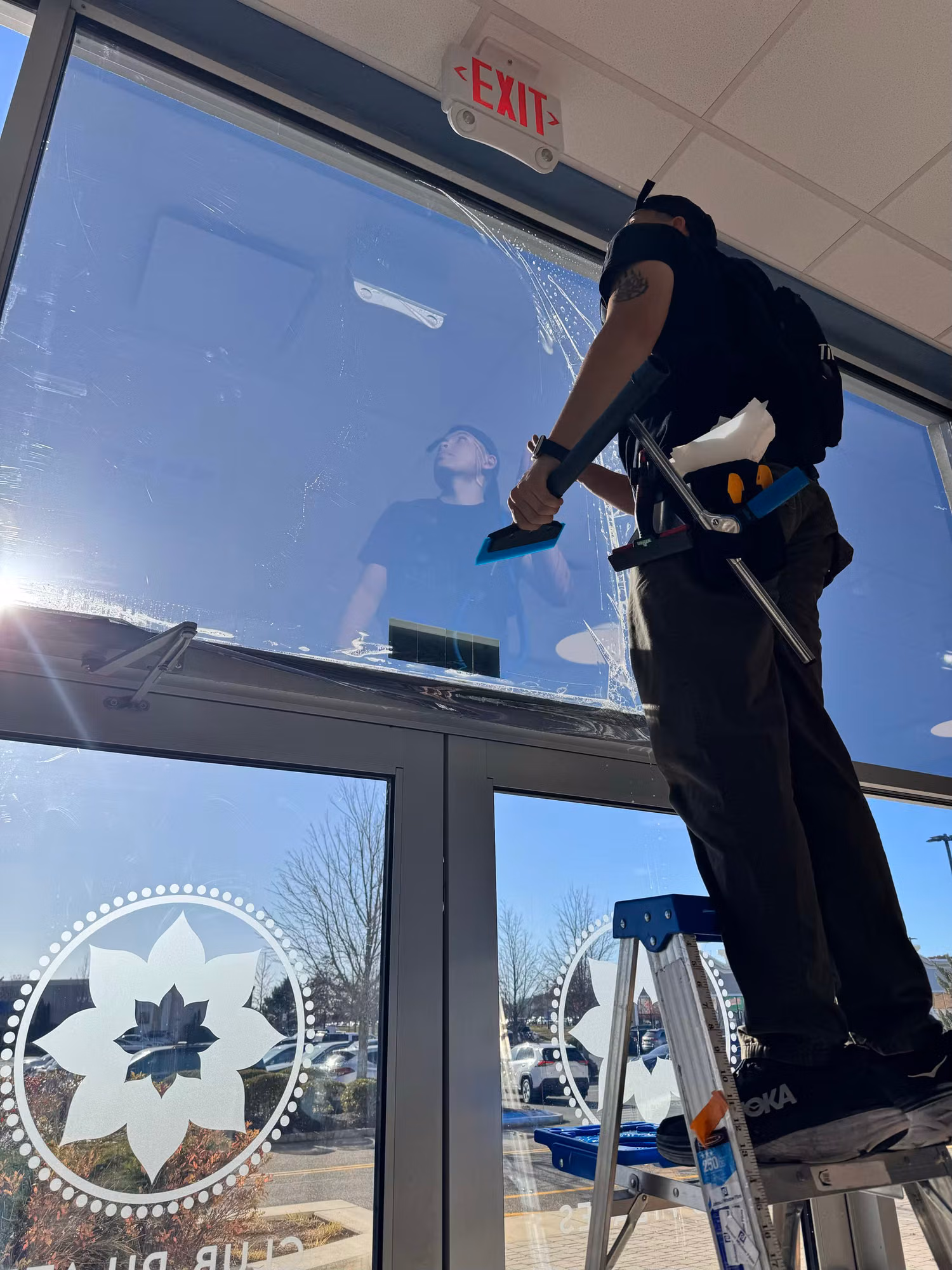 Installer on a ladder squeegeeing window film onto glass entry doors with frosted logo graphics below.