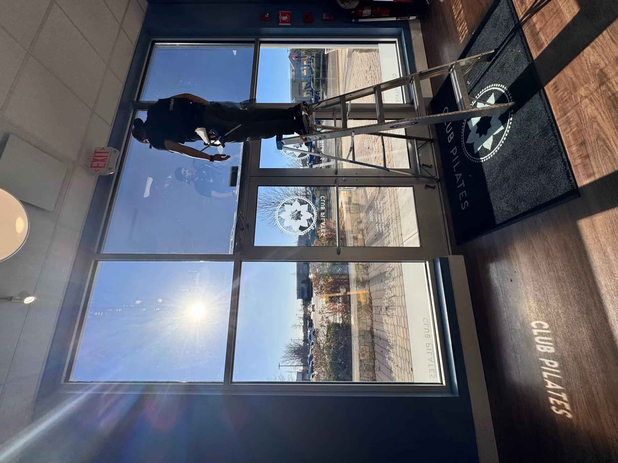 Installer on a ladder applying frosted decorative graphics to floor-to-ceiling storefront glass at a fitness studio entry.