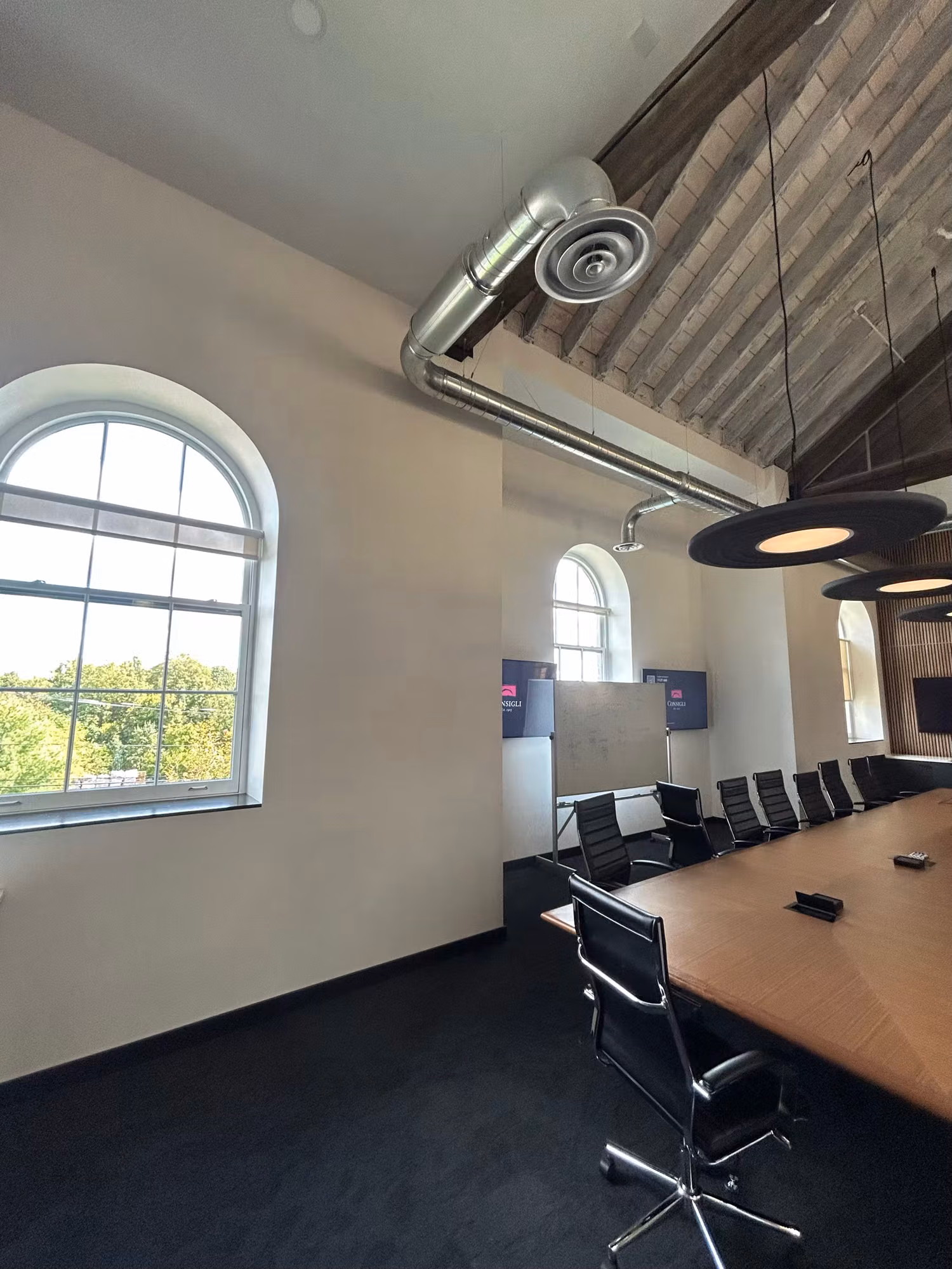 Conference room with tall arched exterior windows, exposed wood ceiling and ductwork, and a long table with black chairs.