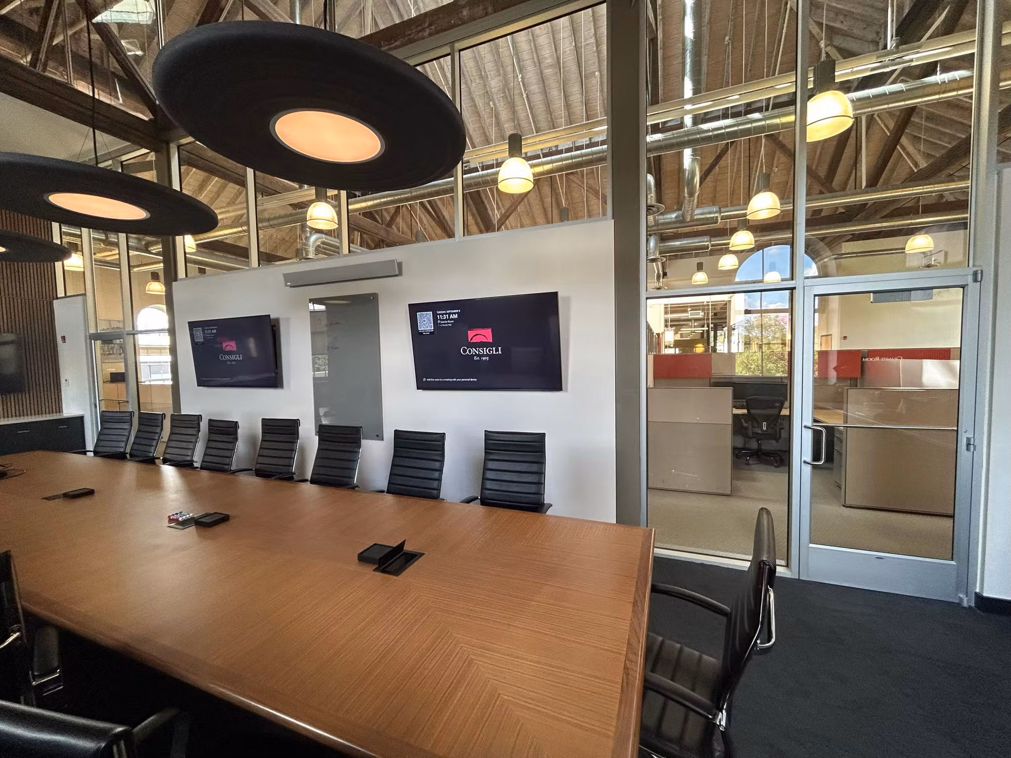 Conference room with a wood table, black chairs, and a floor-to-ceiling glass partition wall and door separating an adjacent office.