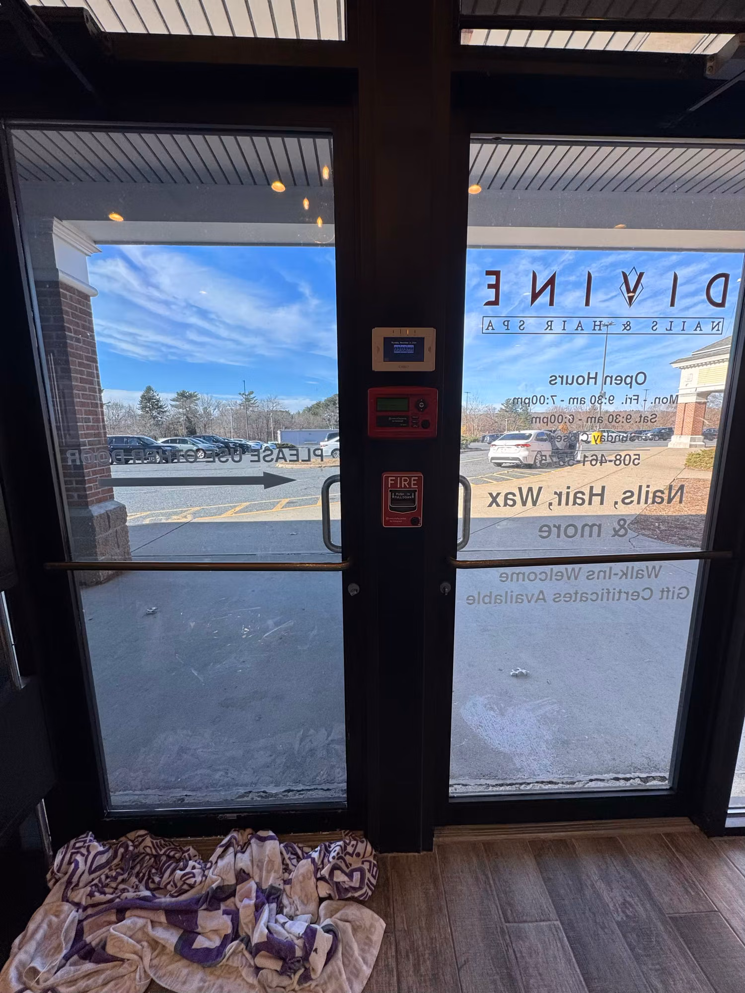Interior view through a glass entry door showing reverse-reading vinyl logo and hours with a parking lot outside.