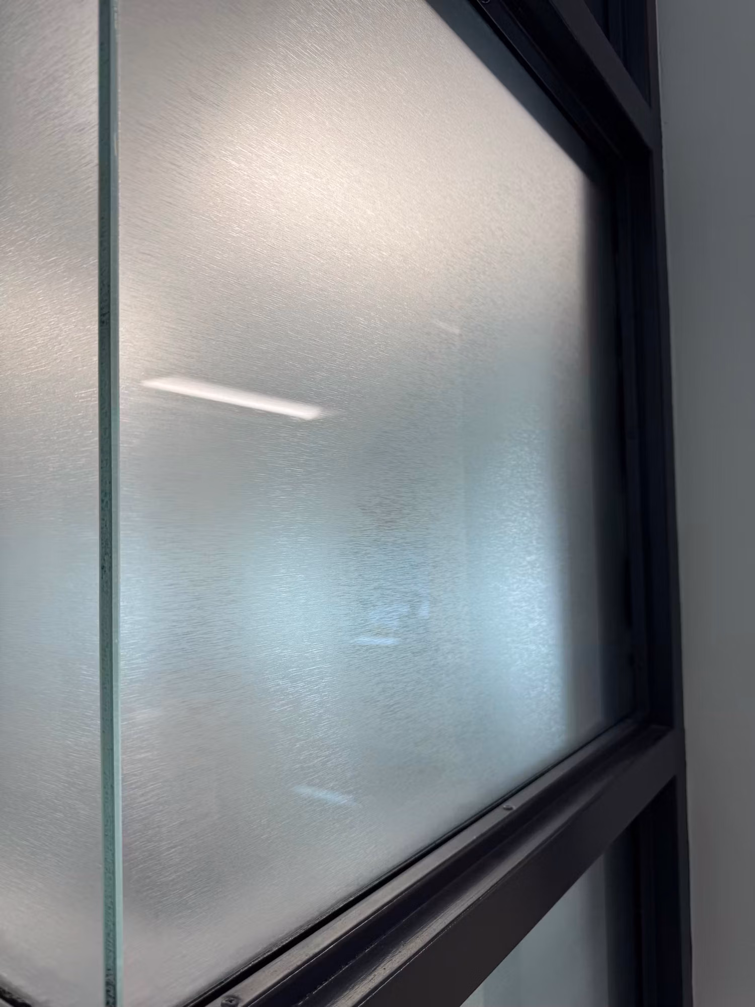 Close-up of a black-framed glass partition pane finished with frosted privacy film diffusing the interior light.