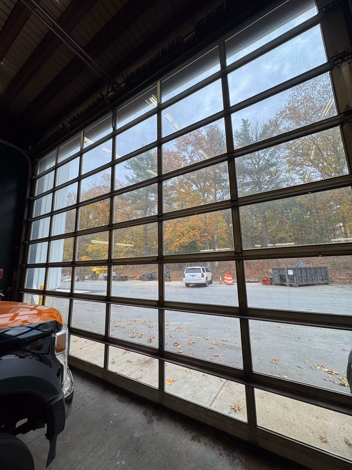 Large full-view glass overhead garage door with multiple panes overlooking a parking lot and trees.