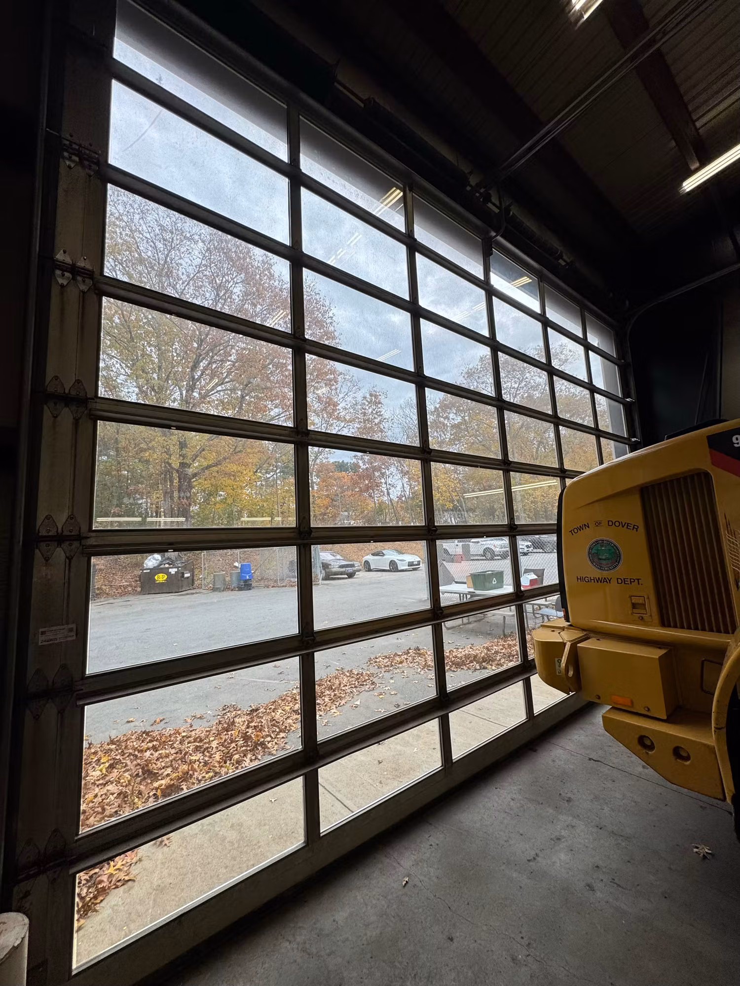 Full-view sectional glass garage door with gridded panes looking out onto a paved lot with parked vehicles.