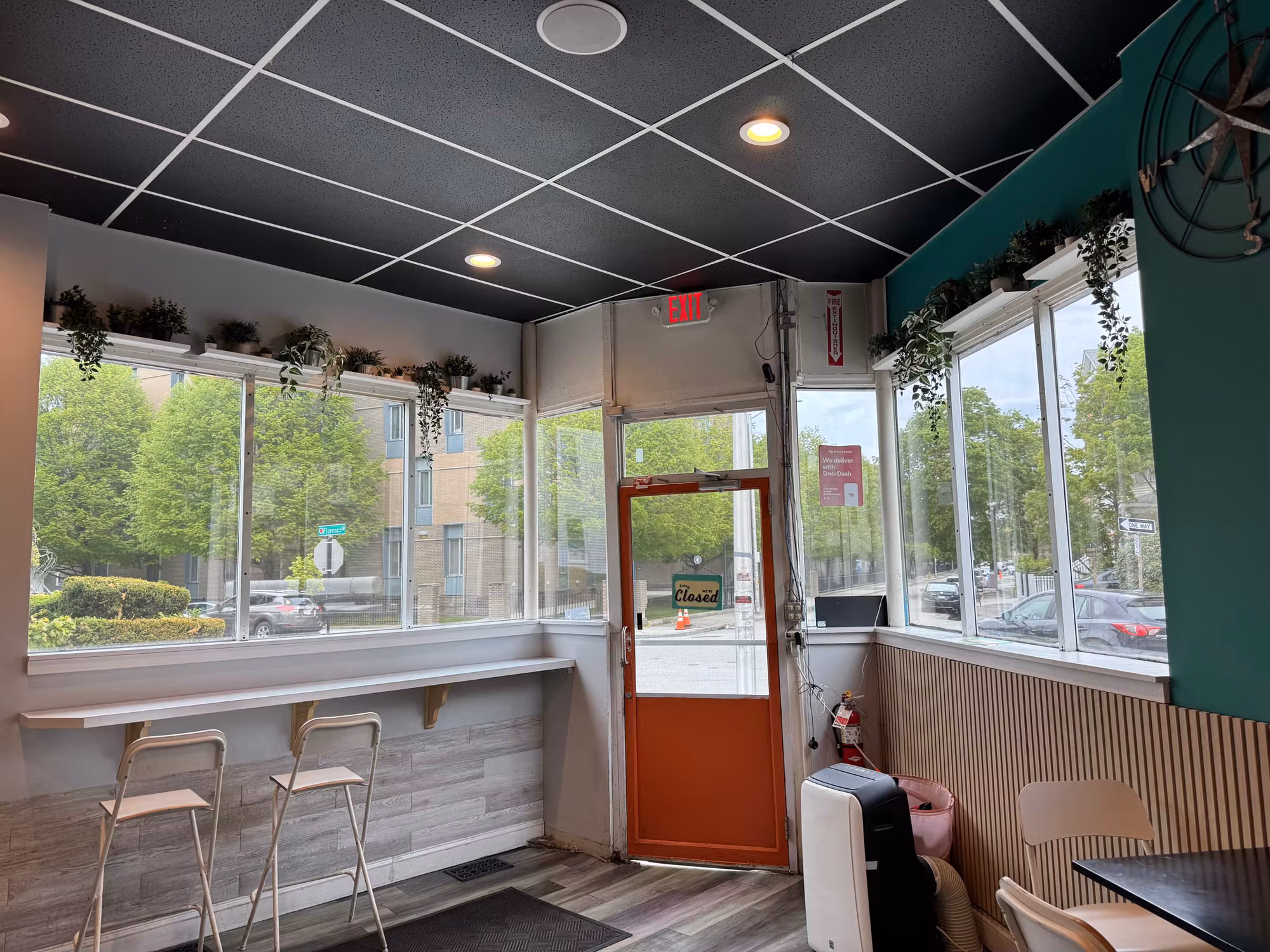 Interior of a casual eatery with wraparound storefront windows and a glass entry door beside bar seating.