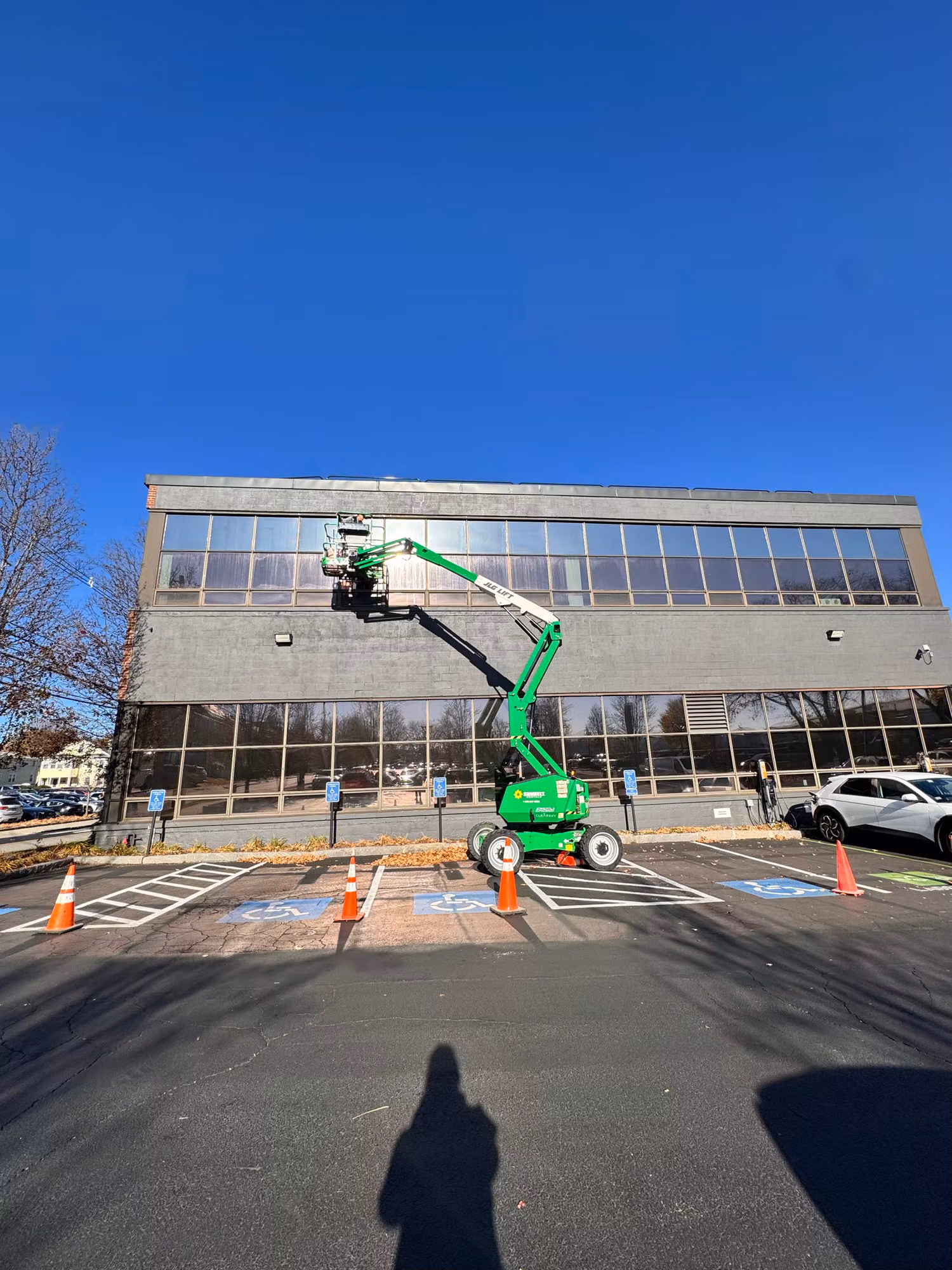 Technician on a green boom lift installing window film across the upper windows of a two-story office building.