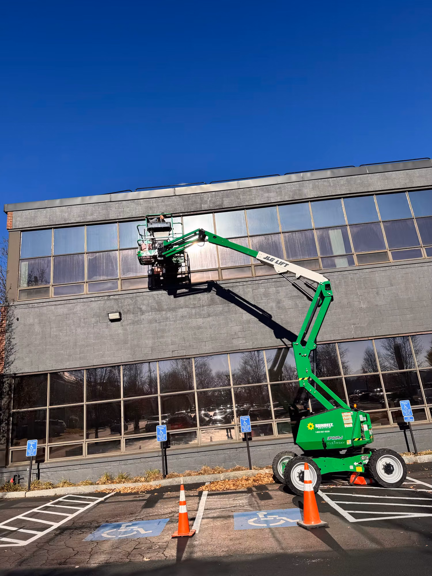 Boom lift raised against an office building facade as a worker applies window film to the second-floor glass.