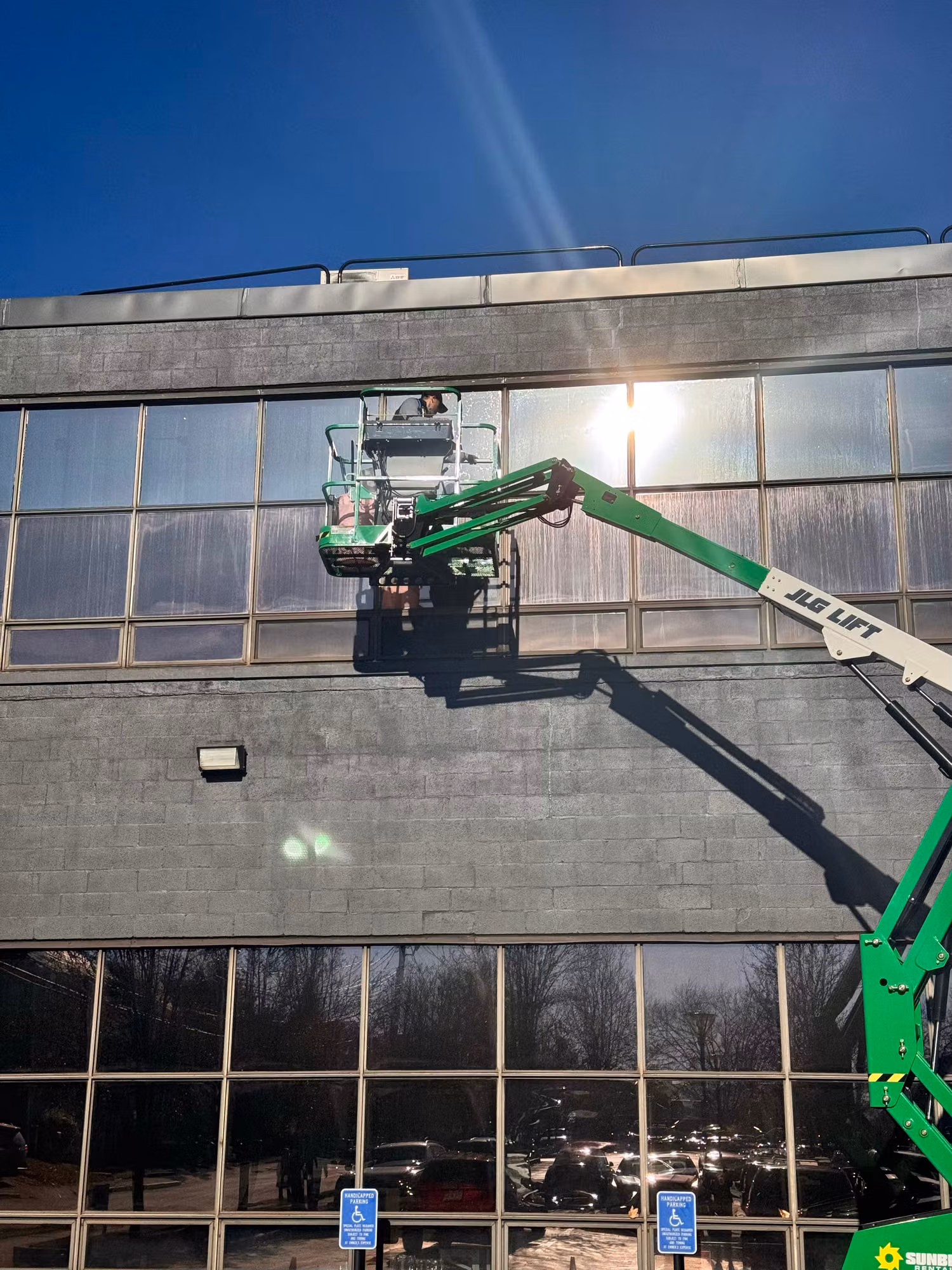 Close view of a worker on a boom lift fitting window film to a strip of second-floor commercial windows in bright sun.