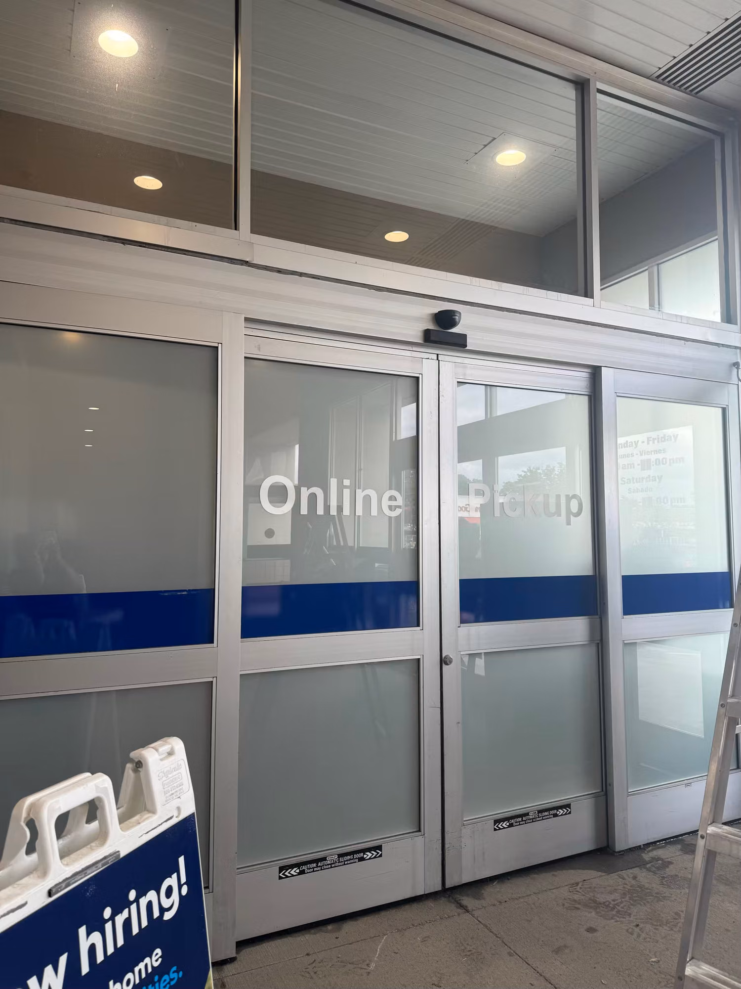 Exterior of automatic entry doors and transom windows with Online Pickup graphics during a film installation, ladder in frame.