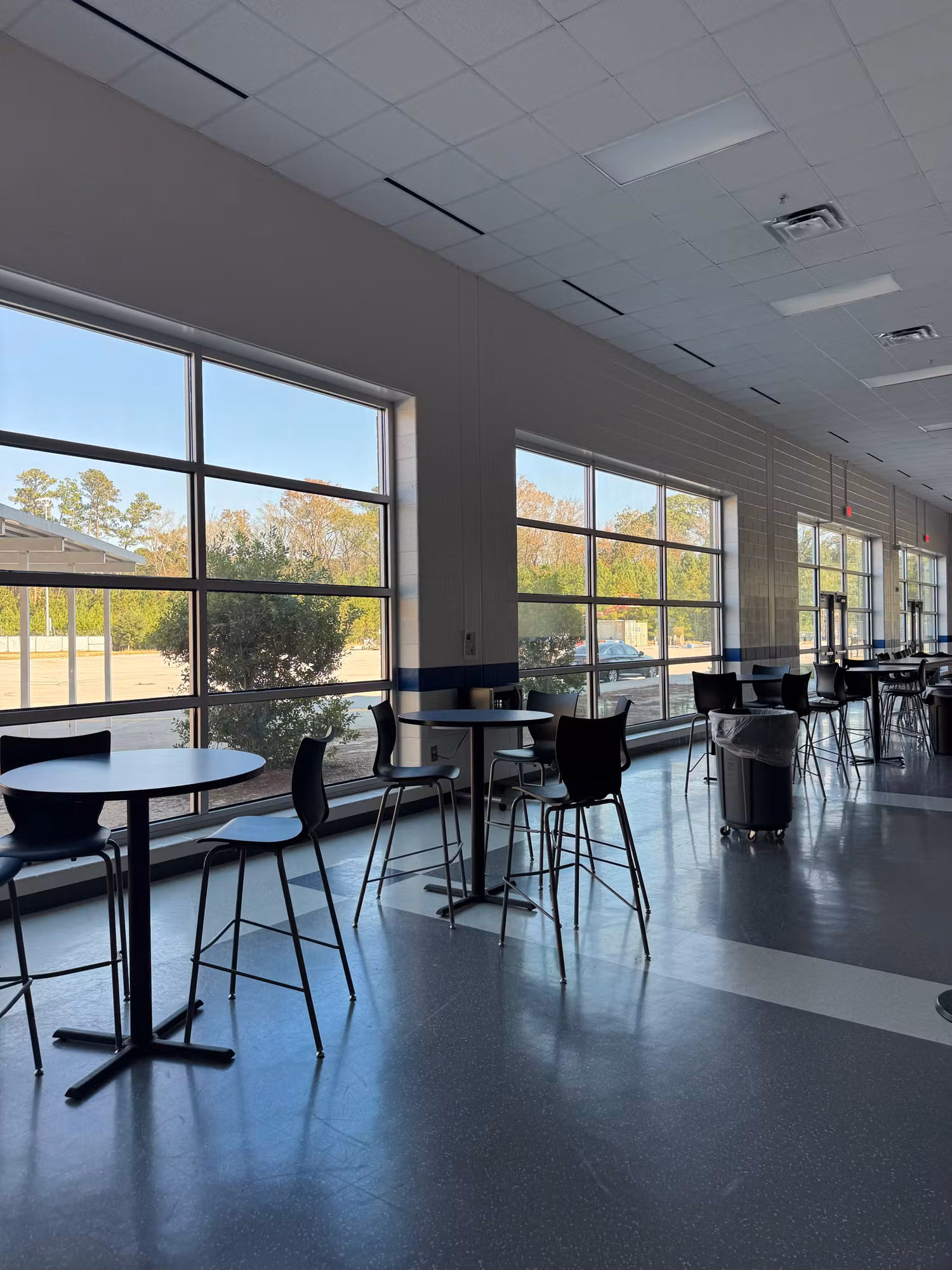 Bright school commons with tall tinted window walls and high-top tables on a polished floor.