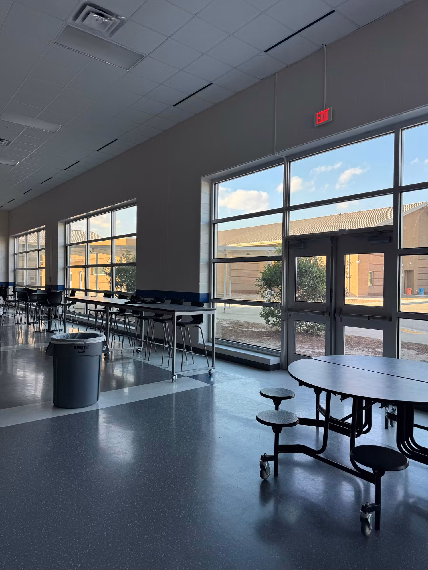 Long tinted window wall and entry door lining a school cafeteria with rows of tables.