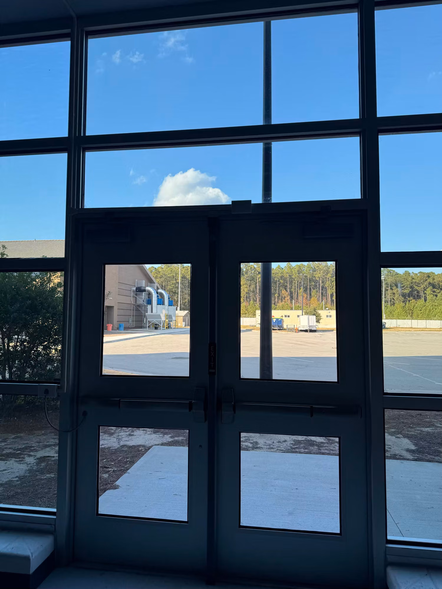 Interior view through a tinted curtain wall and double entry doors looking out toward a parking area.