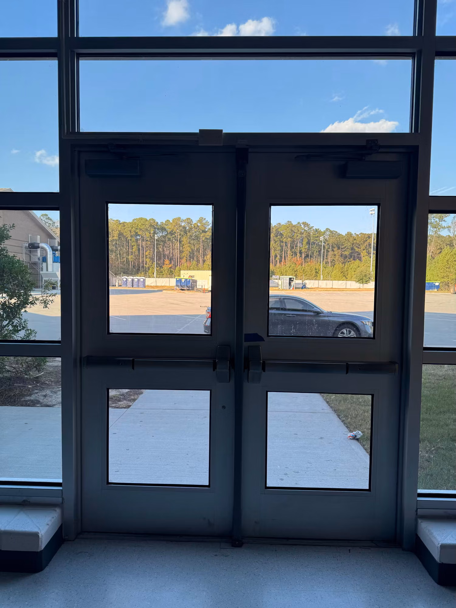 Tinted glass double entry doors set in a window wall with vehicles and trees visible outside.