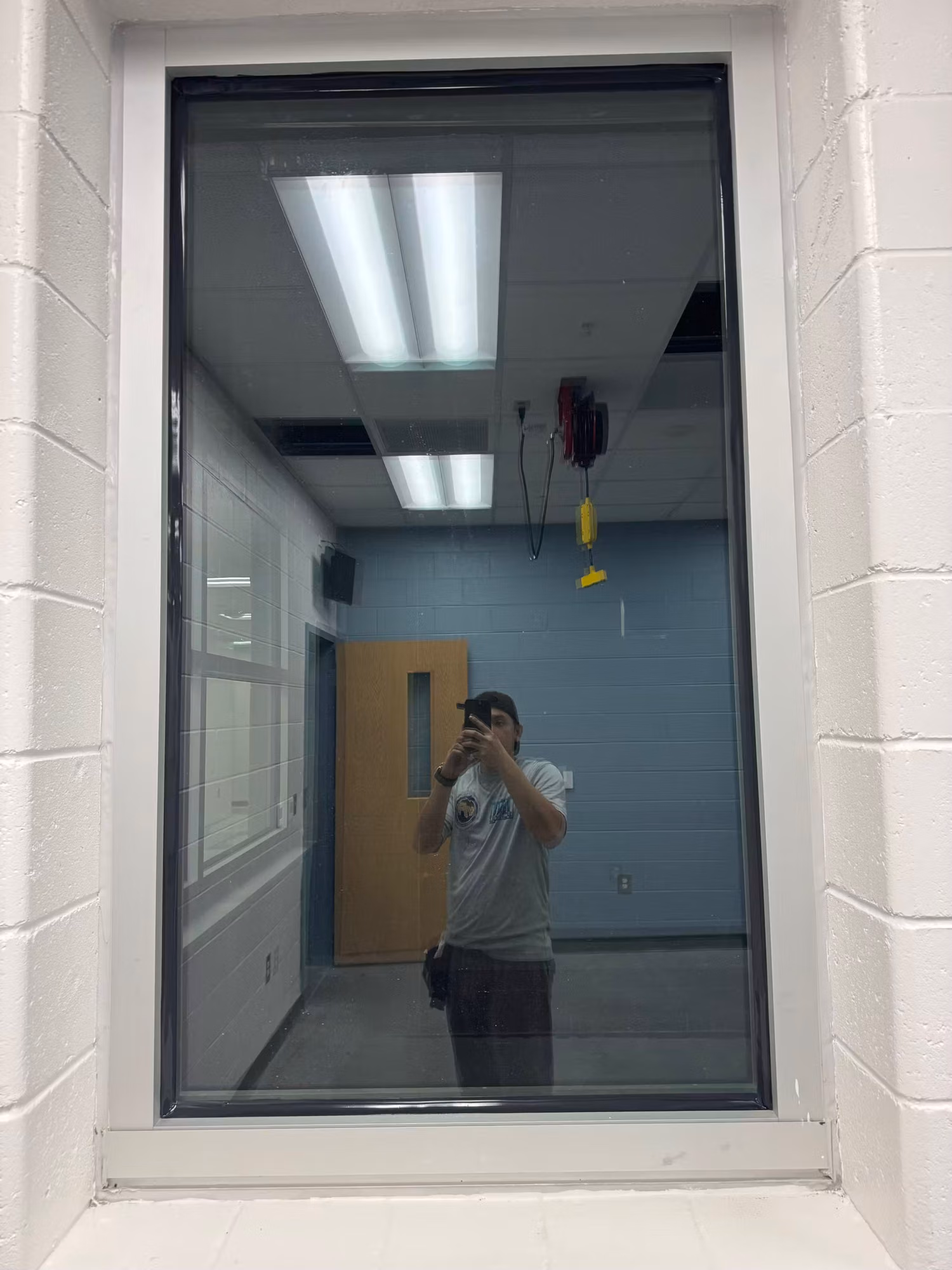 Interior block-wall window with reflective film revealing a corridor and wood door beyond the glass.