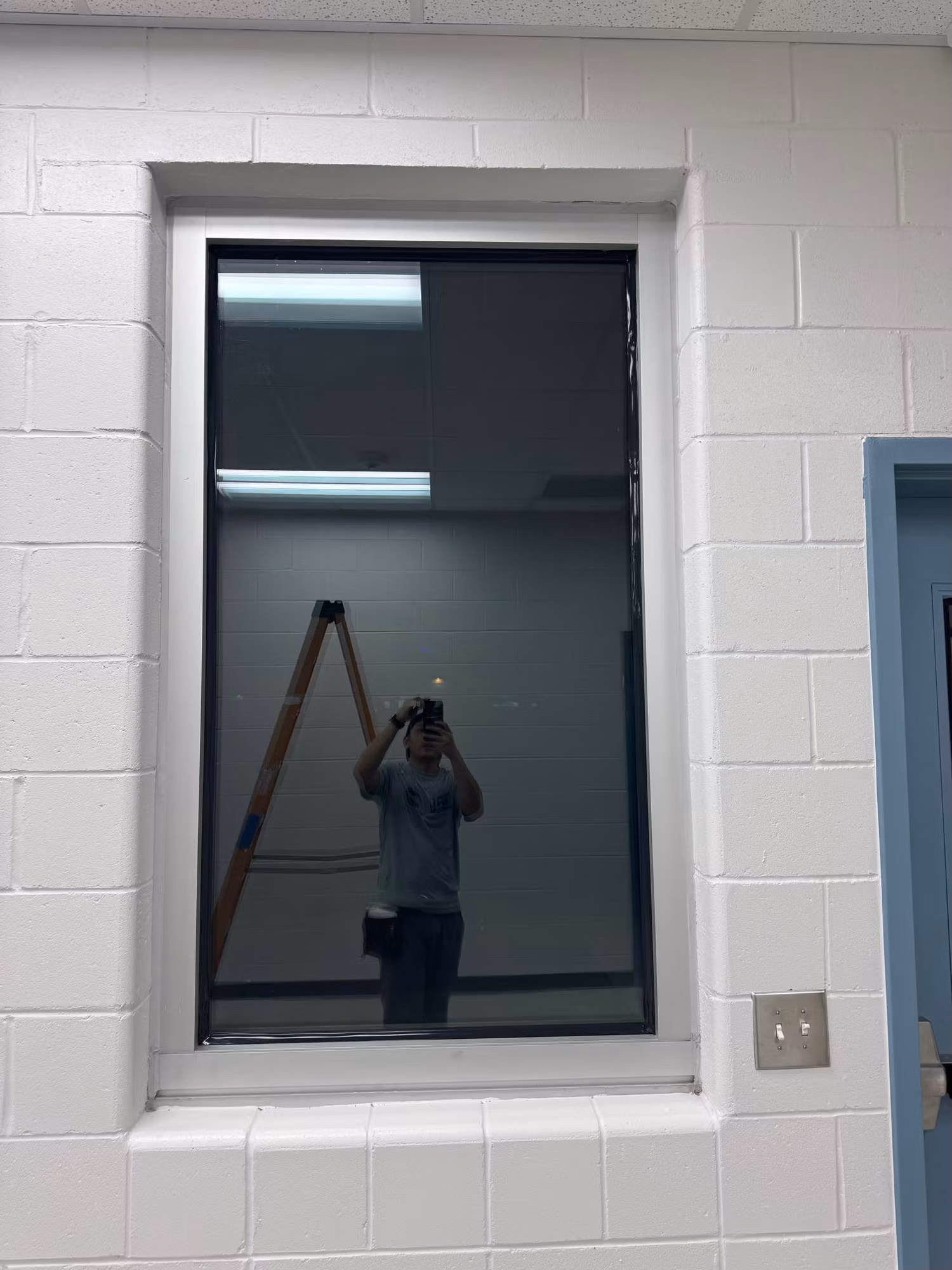 Interior block-wall window with darkened reflective film showing a ladder and work area through the glass.