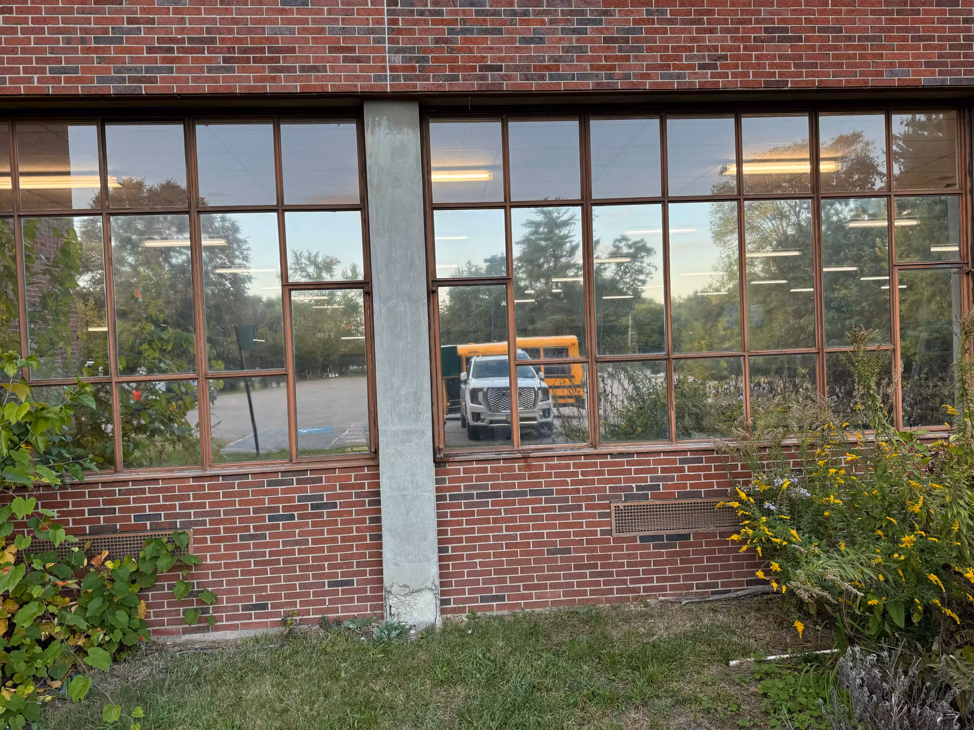 Gridded brick-framed school windows with reflective solar film mirroring trees, sky and a parked vehicle.