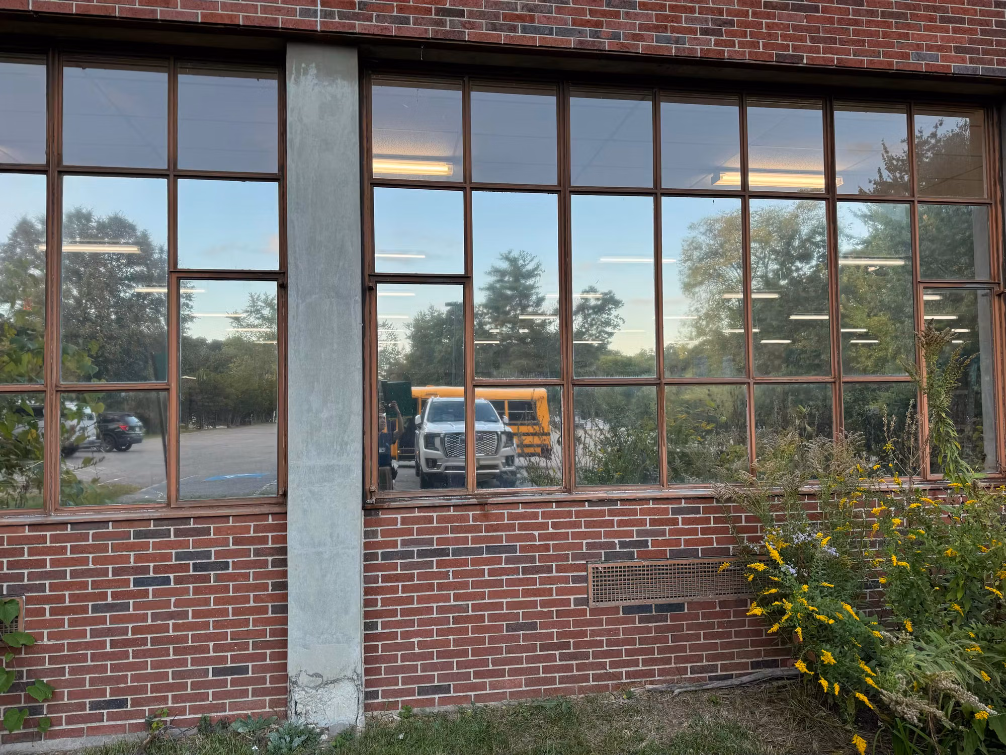 Large multi-pane school window with reflective film showing a parking lot, pickup and bus in the glass.