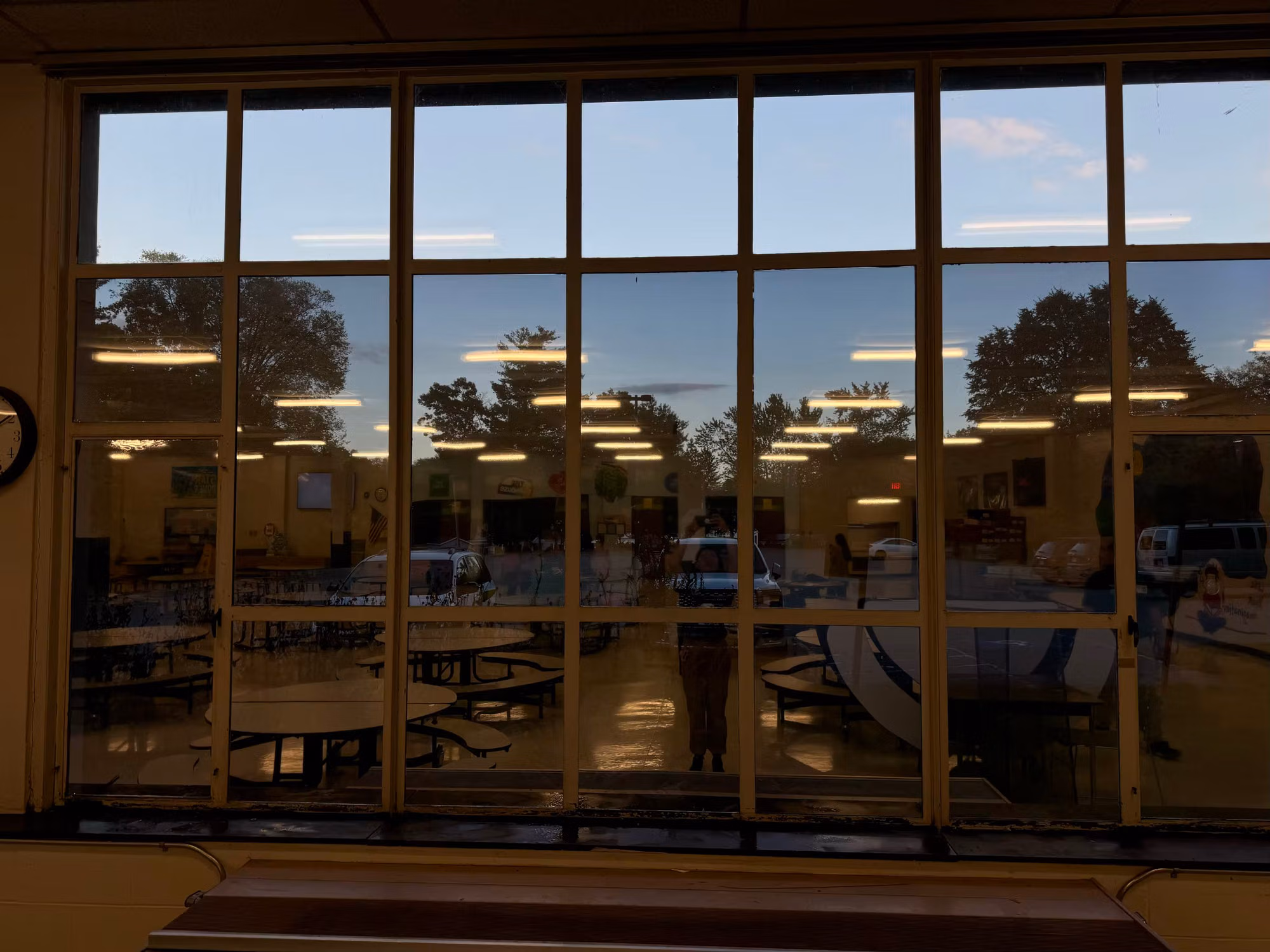 Cafeteria window wall with darkened solar film at dusk, interior lights and tables reflected across the tinted panes.