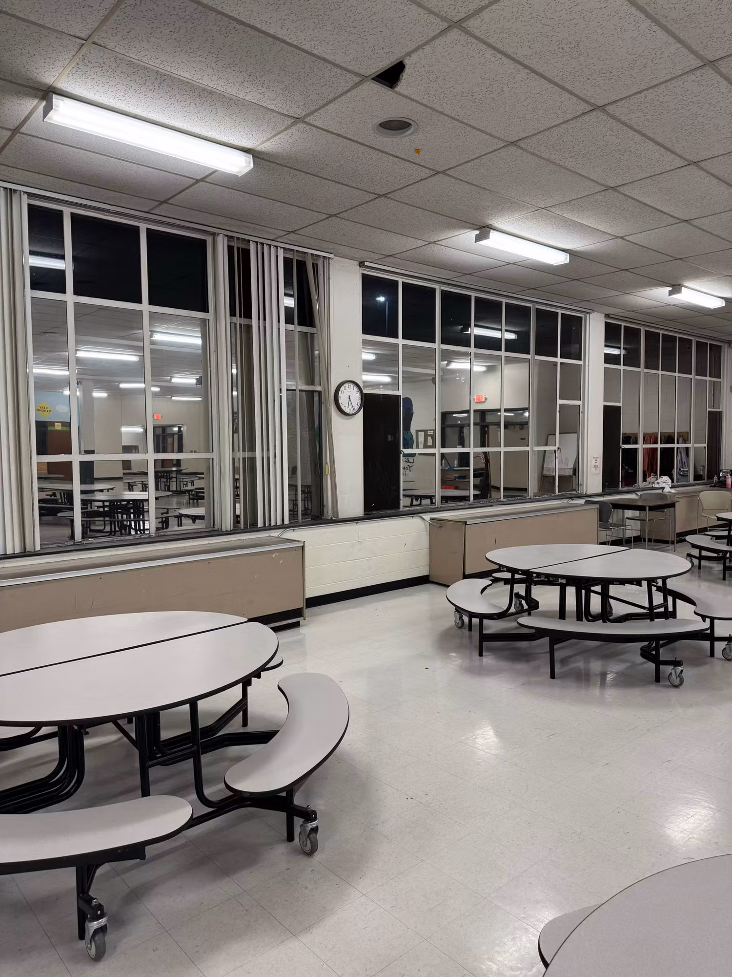 School cafeteria interior at night with a large window wall and wall clock, glass treated with solar film.