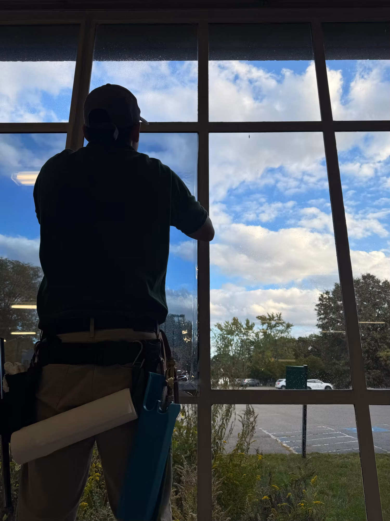 Installer with a tool belt applying window film to a large interior cafeteria window overlooking a parking lot.