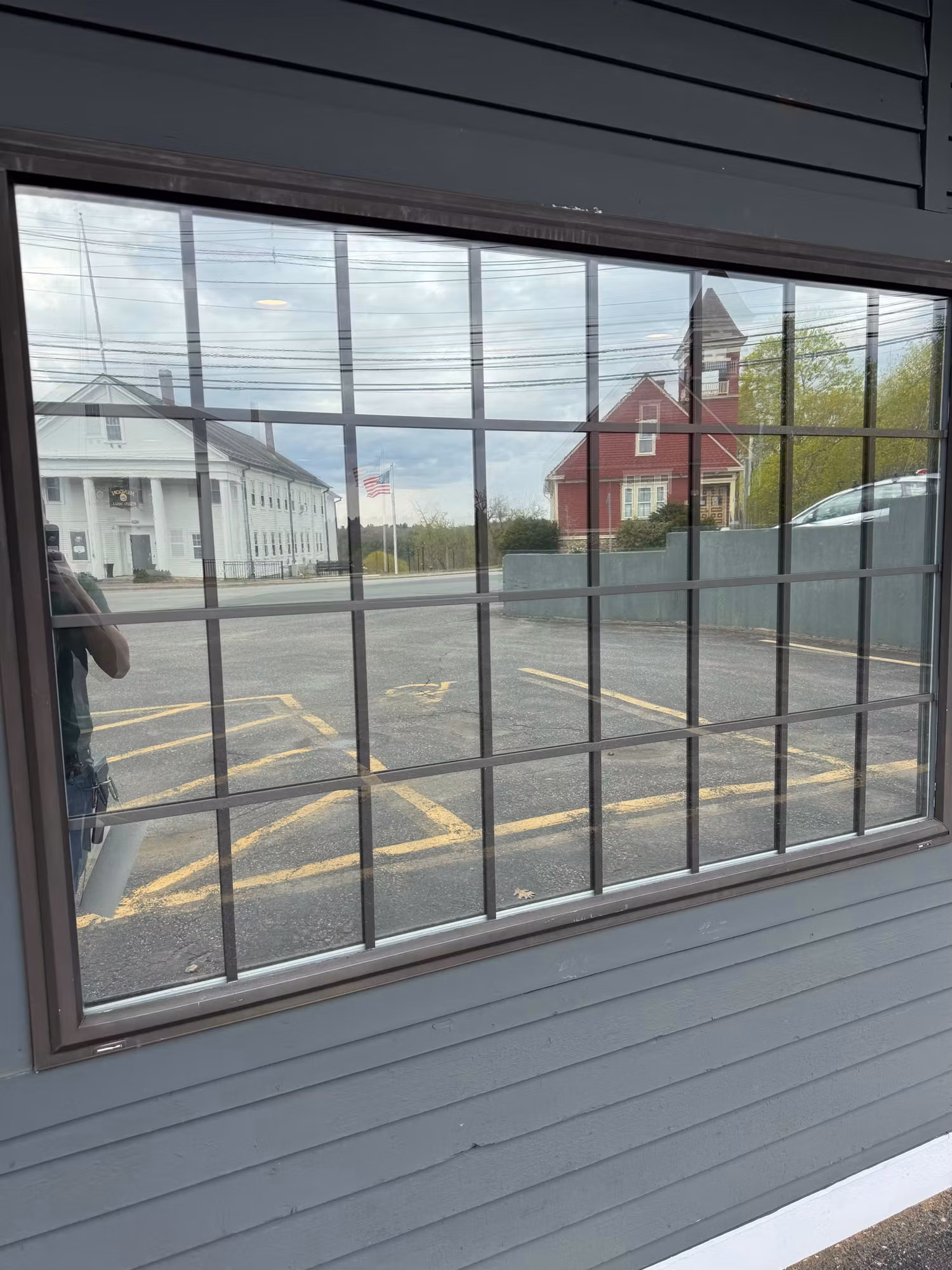 Exterior close-up of a large gridded picture window with darkened glass set into gray clapboard siding facing a parking lot.