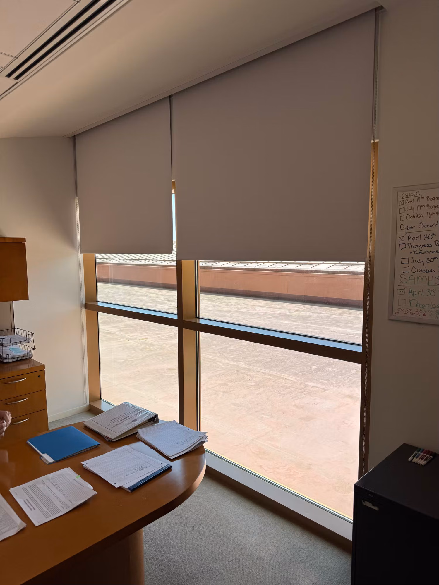 Corner office with two tall windows and grey roller shades partly lowered above a wood desk stacked with paperwork.