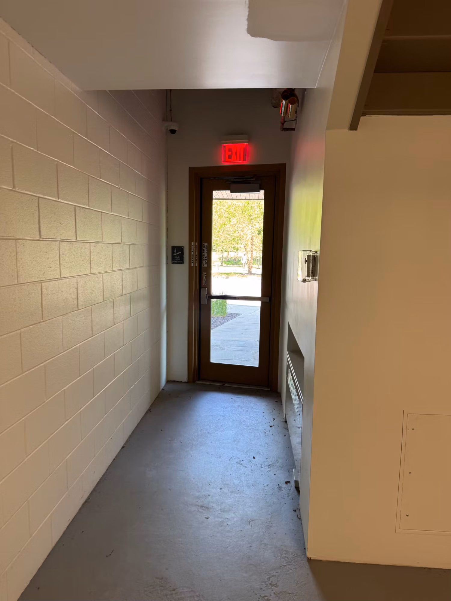 Glass exit door at the end of a block-walled corridor letting daylight into the concrete hallway.