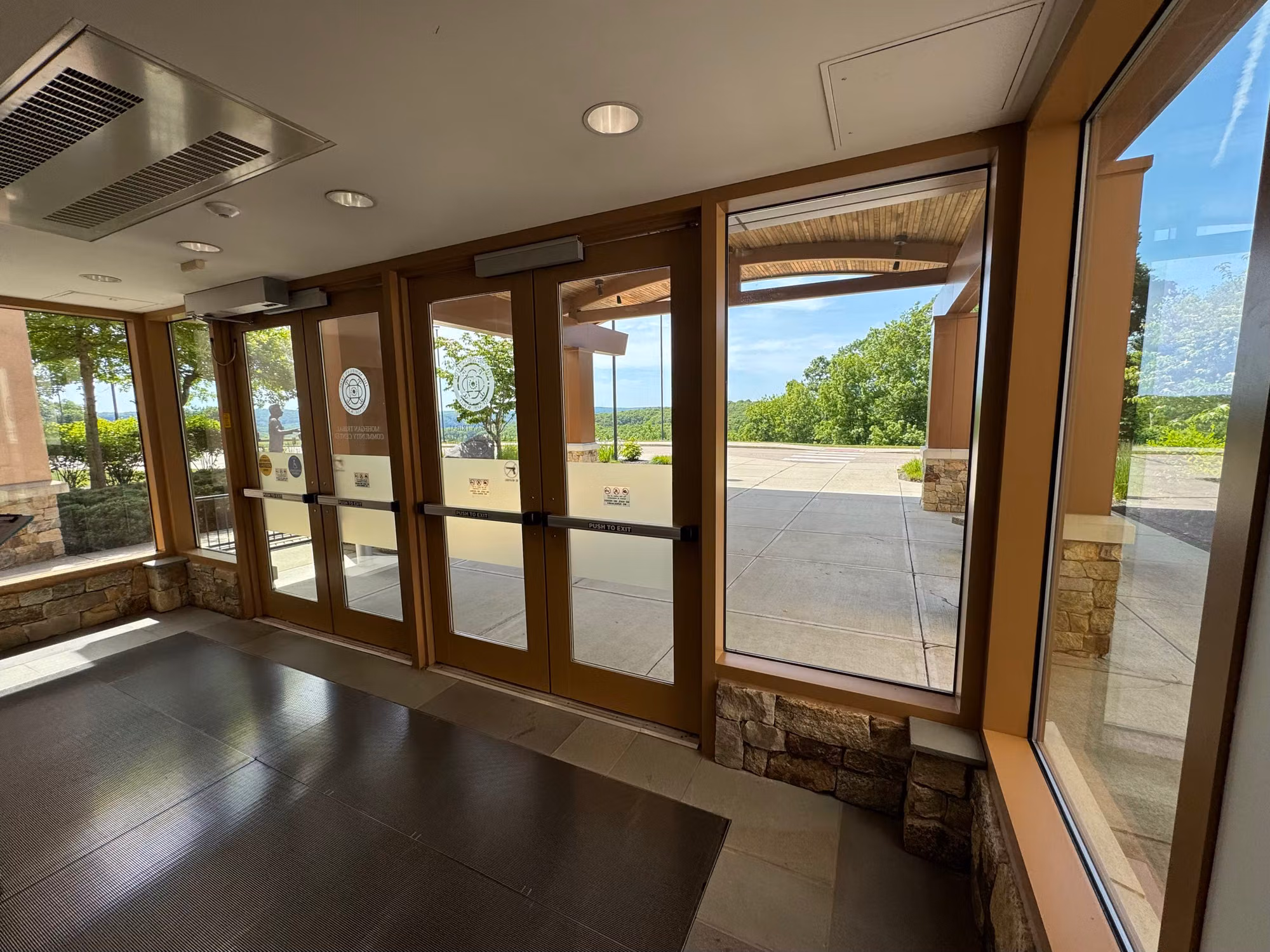 Building lobby with wood-framed glass entry doors and large picture windows on a stone base under a sunny sky.
