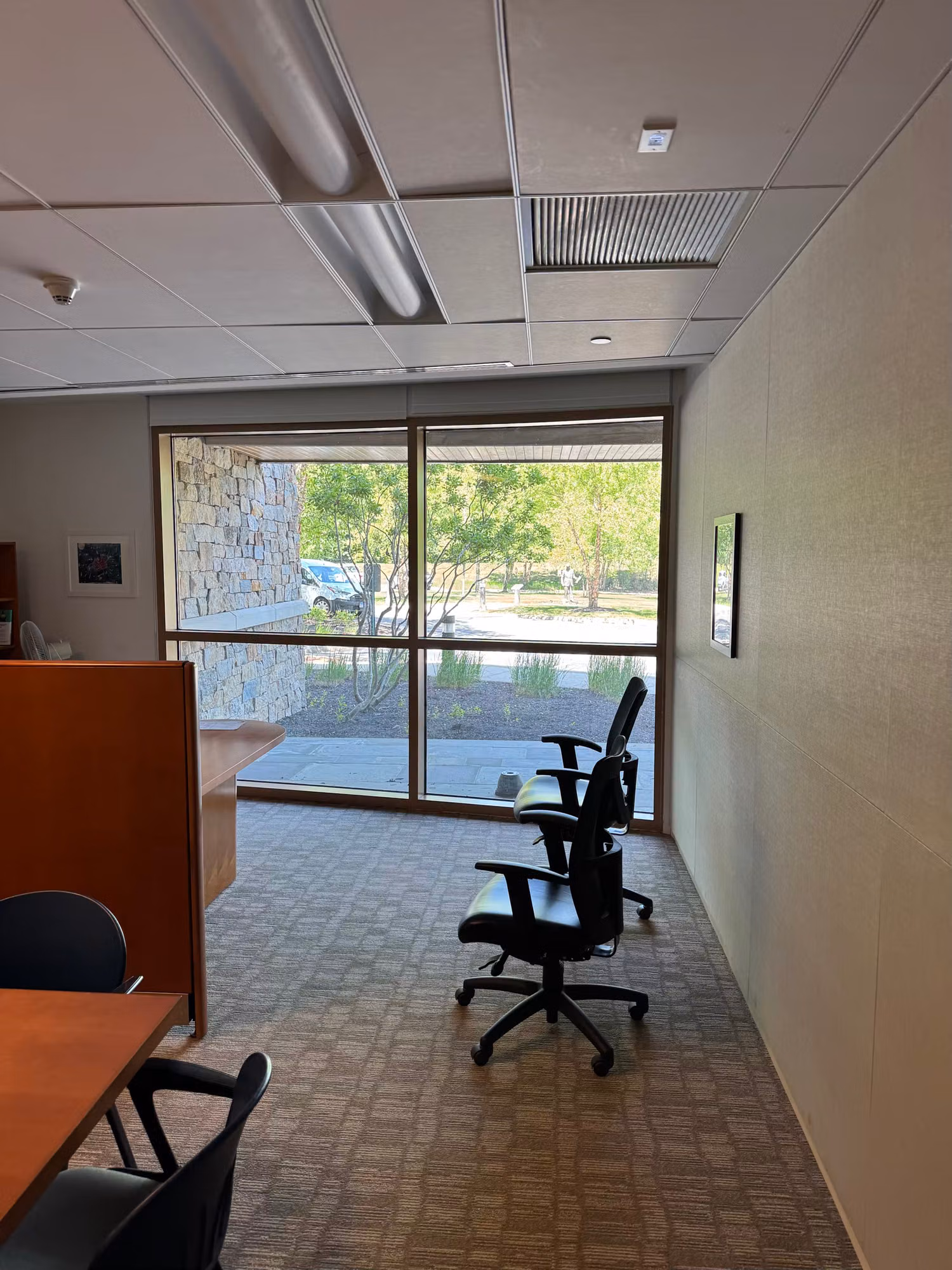 Office window wall of glass overlooking a stone exterior and parking lot beside a desk and task chair.