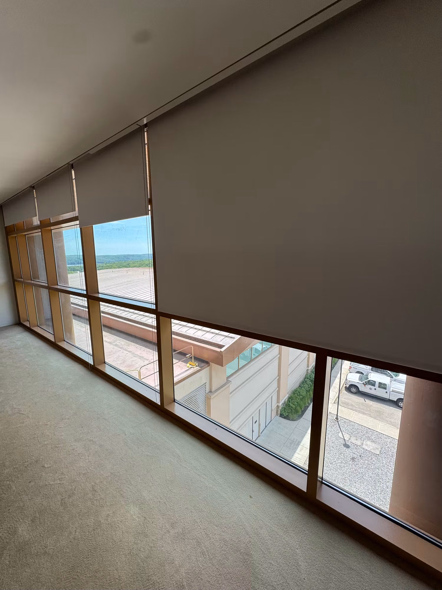 Long run of office windows fitted with grey roller solar shades over glass overlooking the parking area.