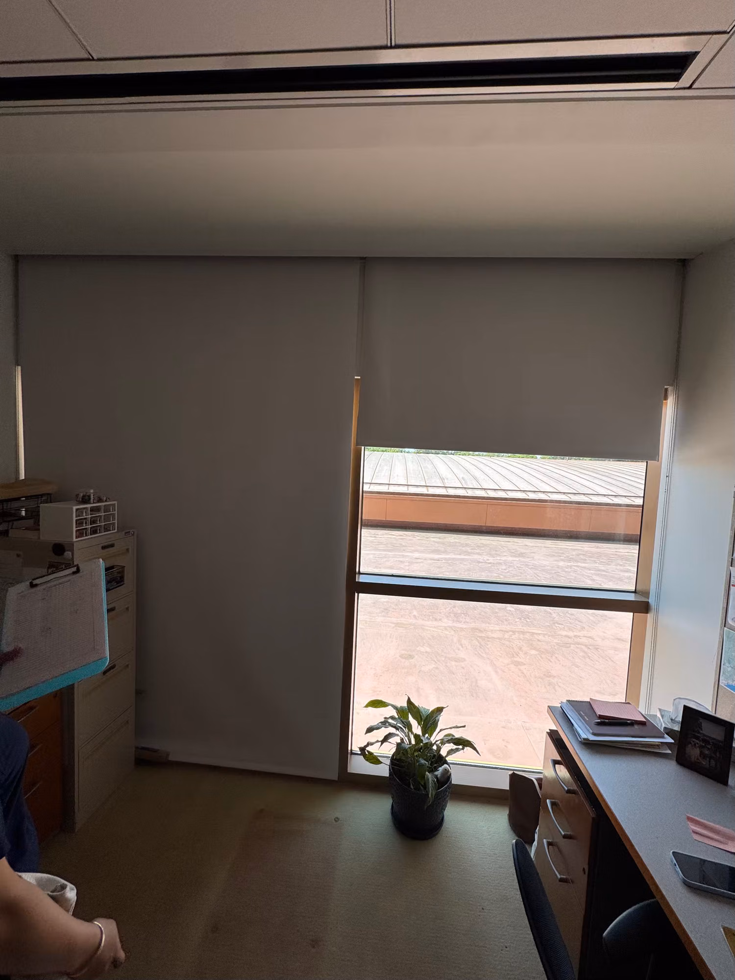 Private office with a tall window covered by a grey roller shade beside a desk and storage cabinet.