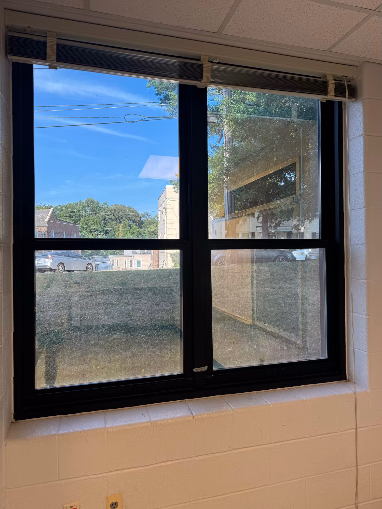 Black-framed sliding classroom window with darkened solar film cutting glare from the field outside.