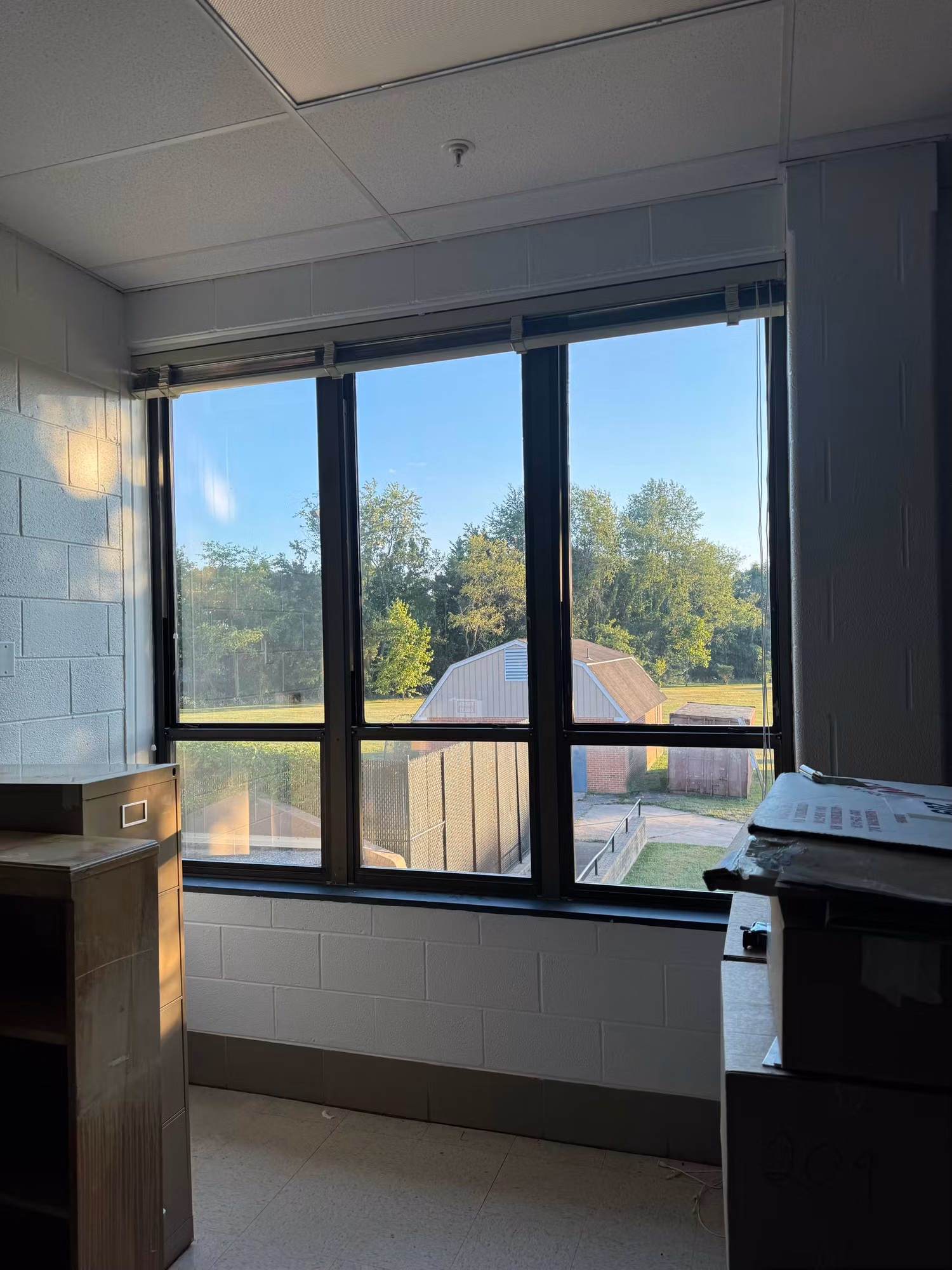 Classroom window looking out over a grassy field and trees beside cinder block walls with a copier nearby.