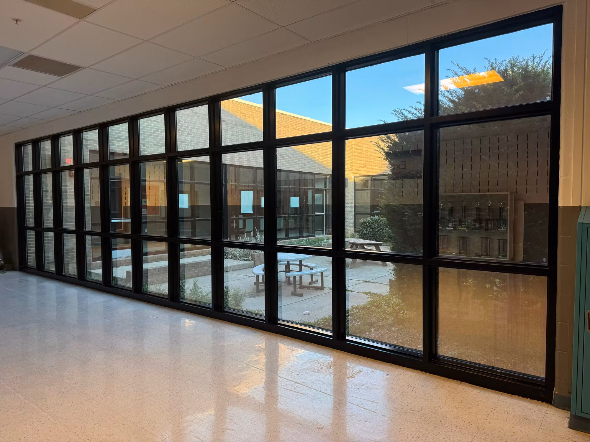 Long school corridor with a floor-to-ceiling glass curtain wall framing a sunlit courtyard and picnic tables.