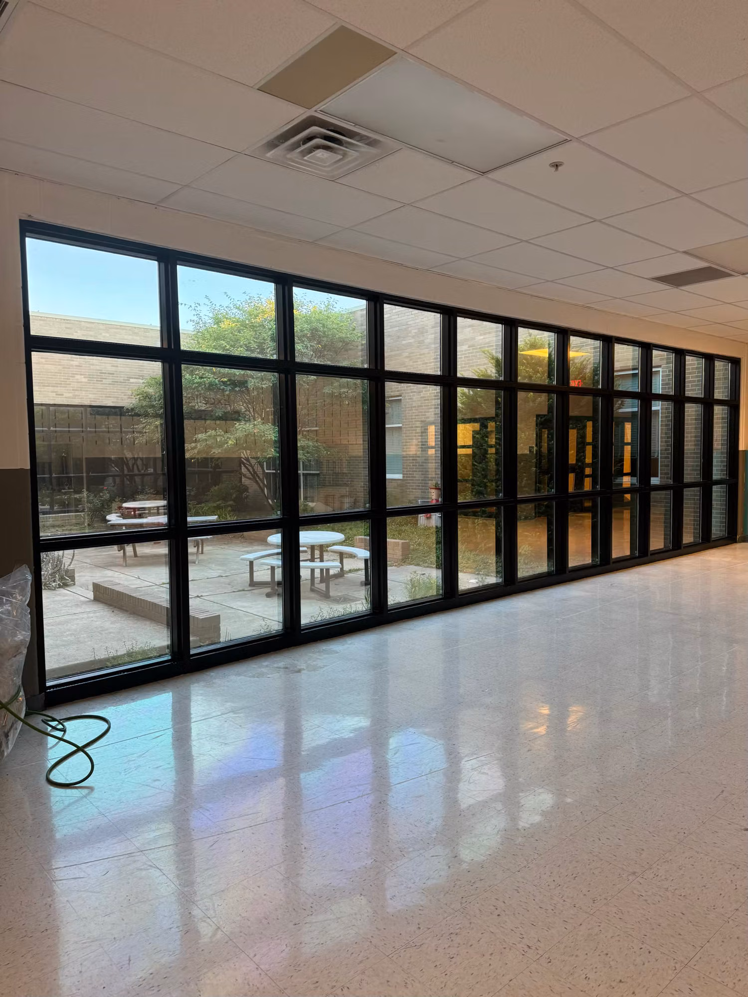 School corridor with a long black-framed glass curtain wall and reflective tile floor facing a courtyard.