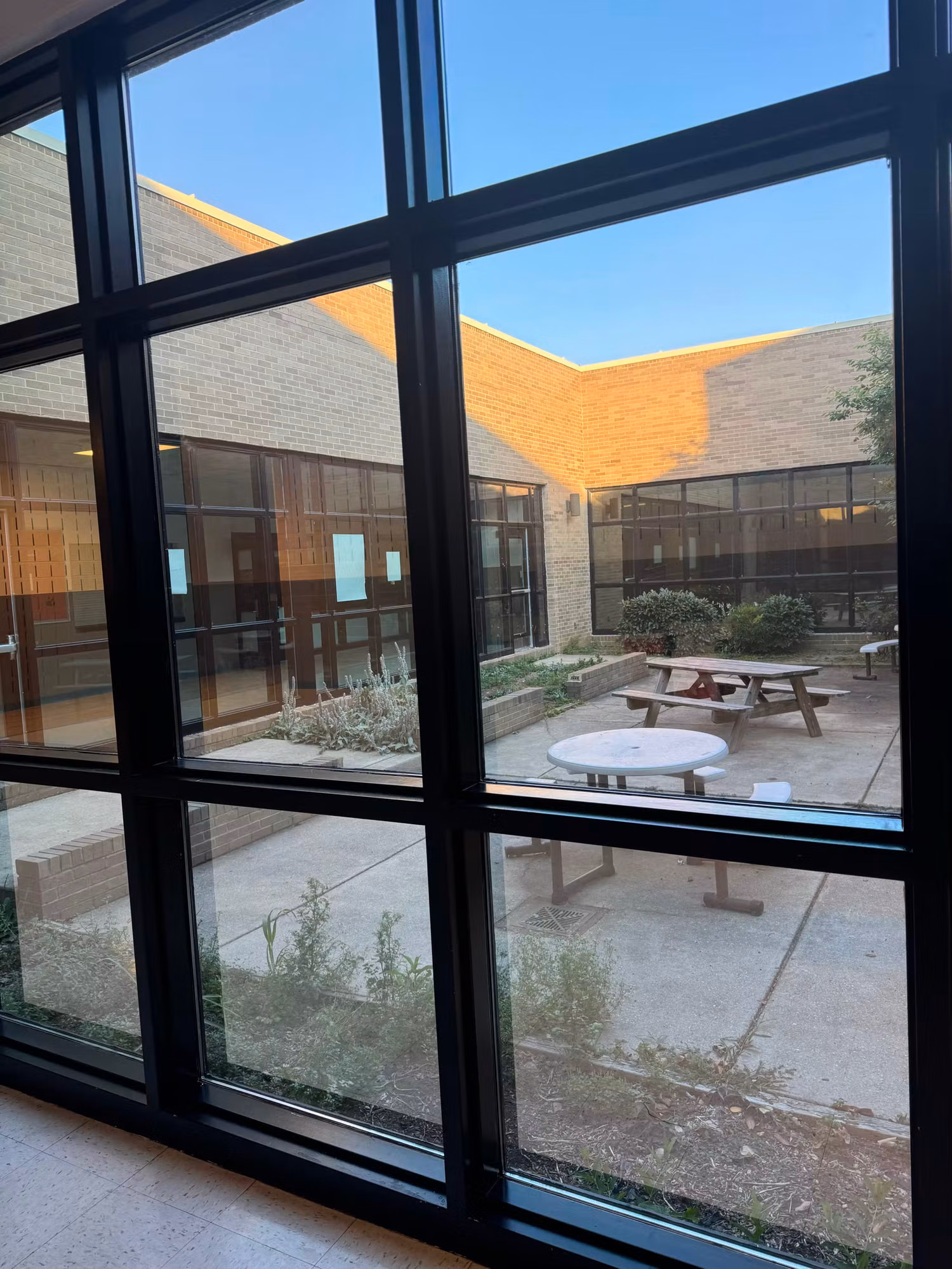 View through a black-framed window grid out to a school courtyard with a picnic table and brick walls.