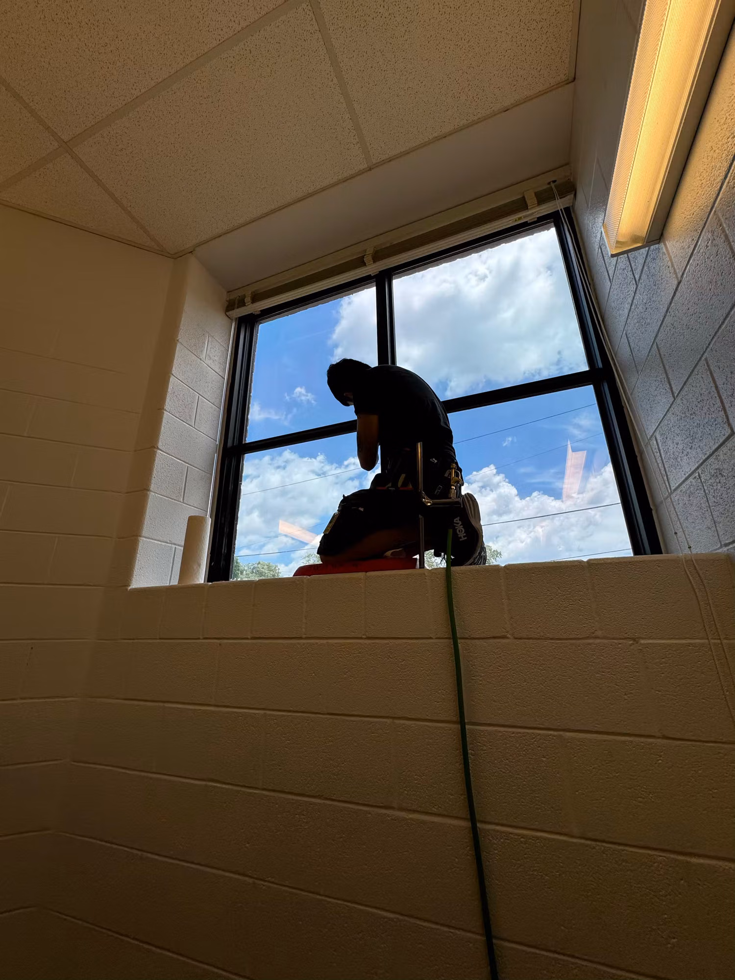 Installer on a lift applying window film to a tall stairwell window set in a block wall against a bright sky.