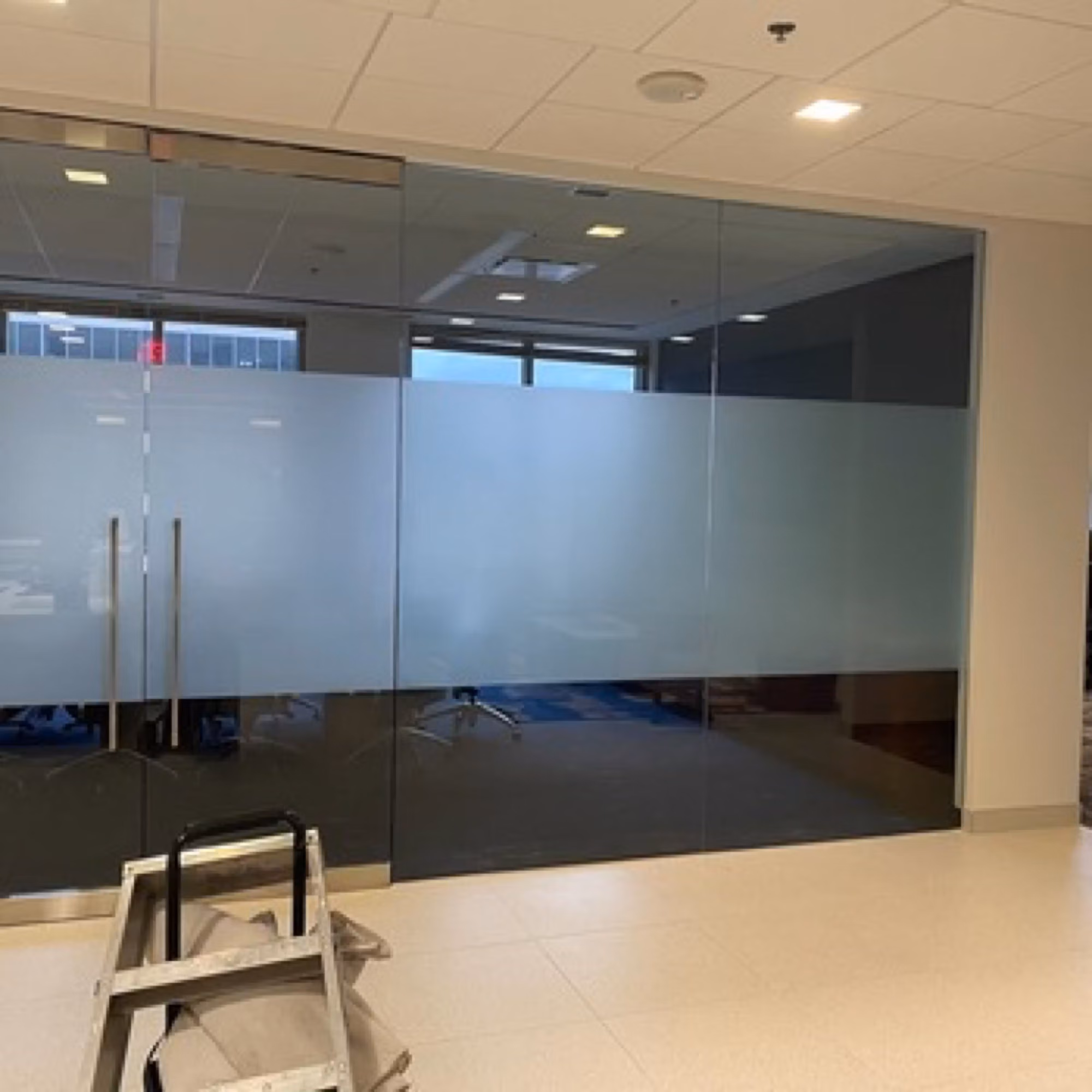 Frameless glass office partition getting a frosted privacy band installed, with a ladder set up during the work.