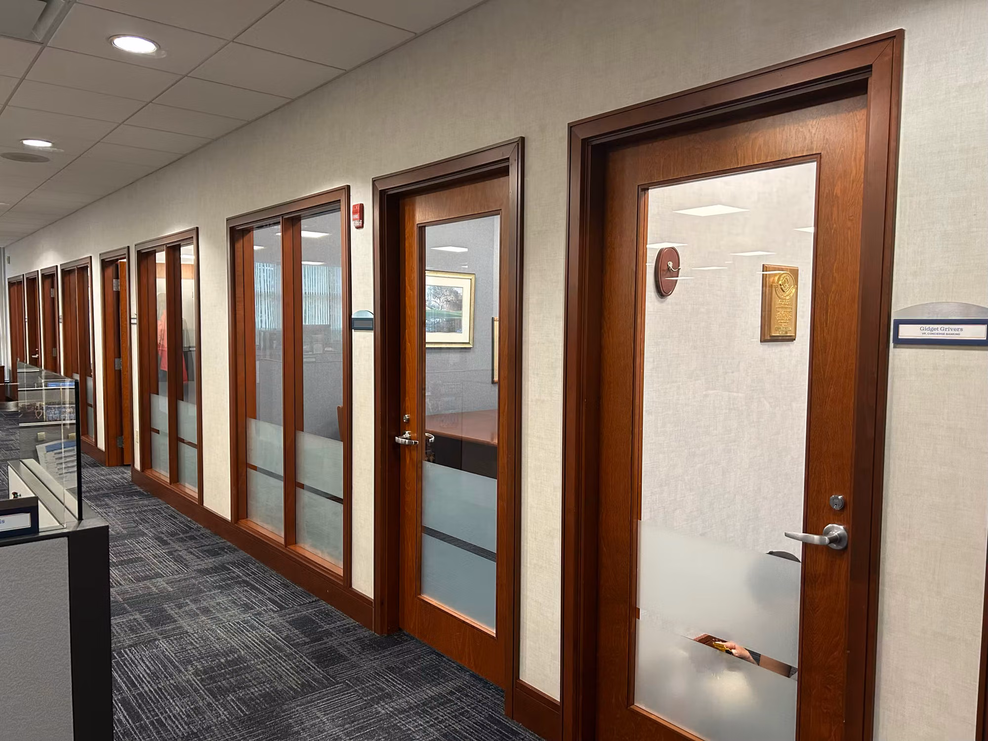 Office corridor lined with wood-framed glass fronts and doors finished with frosted privacy film on the lower glass.