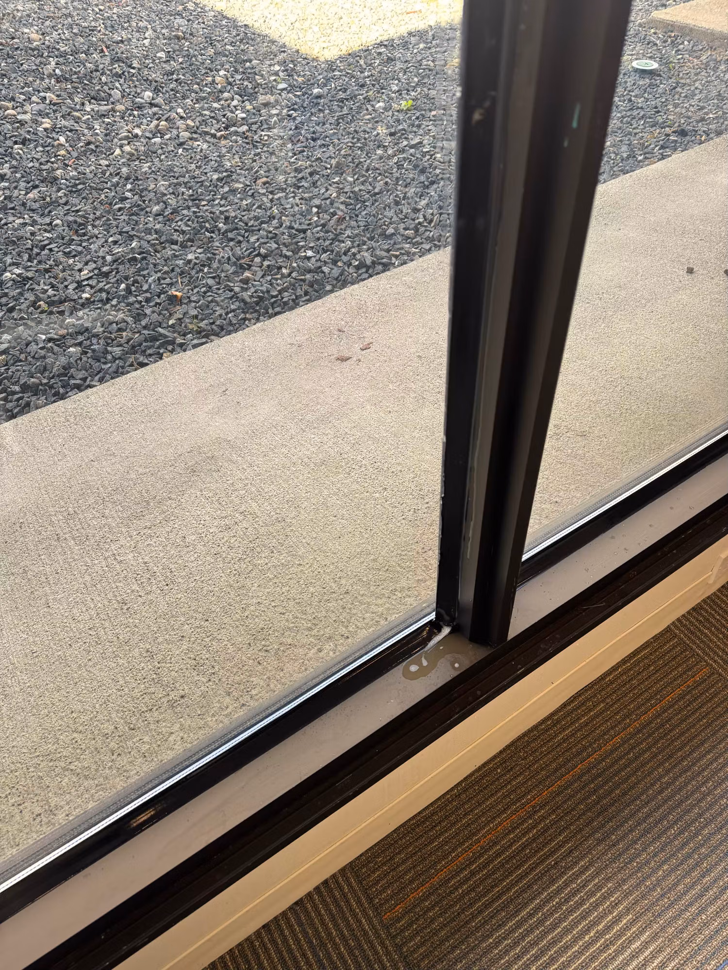 Close-up of the base of a commercial glass door at the floor track beside an entry mat.